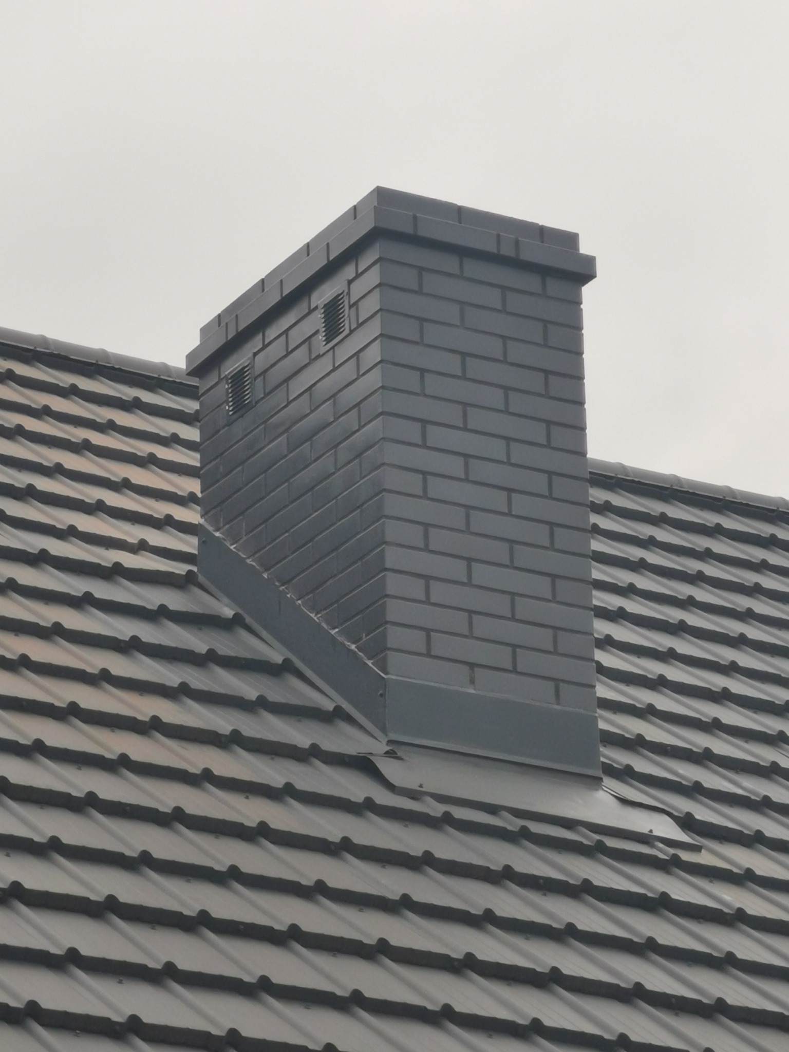 Szary, murowany komin z wentylacją, wystający ponad ciemny dach z dachówki ceramicznej w pochmurny dzień.