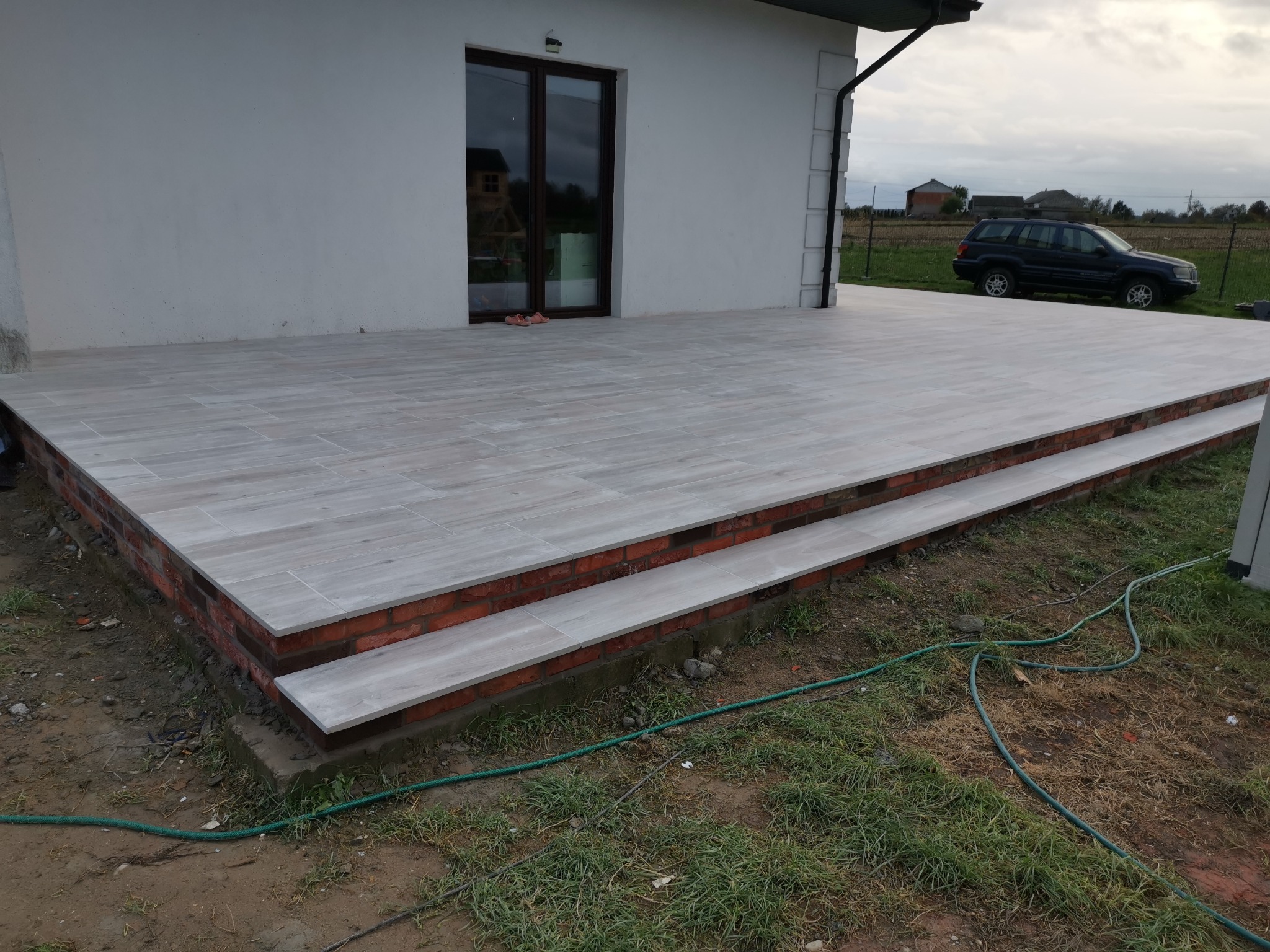 Wyłożony płytkami taras z dwoma stopniami, wykończony czerwoną cegłą, przylegający do białego domu z ciemnymi oknami, na tle zieleni i zaparkowanego samochodu.