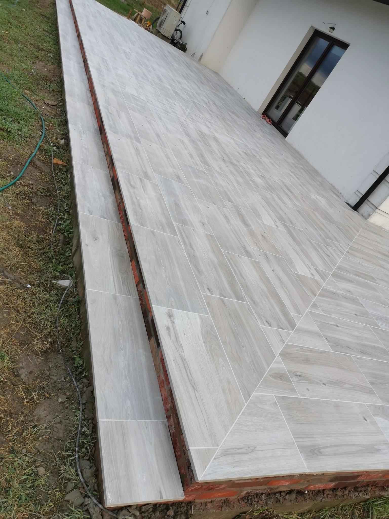 Wyłożony jasnoszarą płytką taras z imitacją drewna, z widoczną ceglaną podmurówką i fragmentem trawnika oraz elewacji domu z oknem.