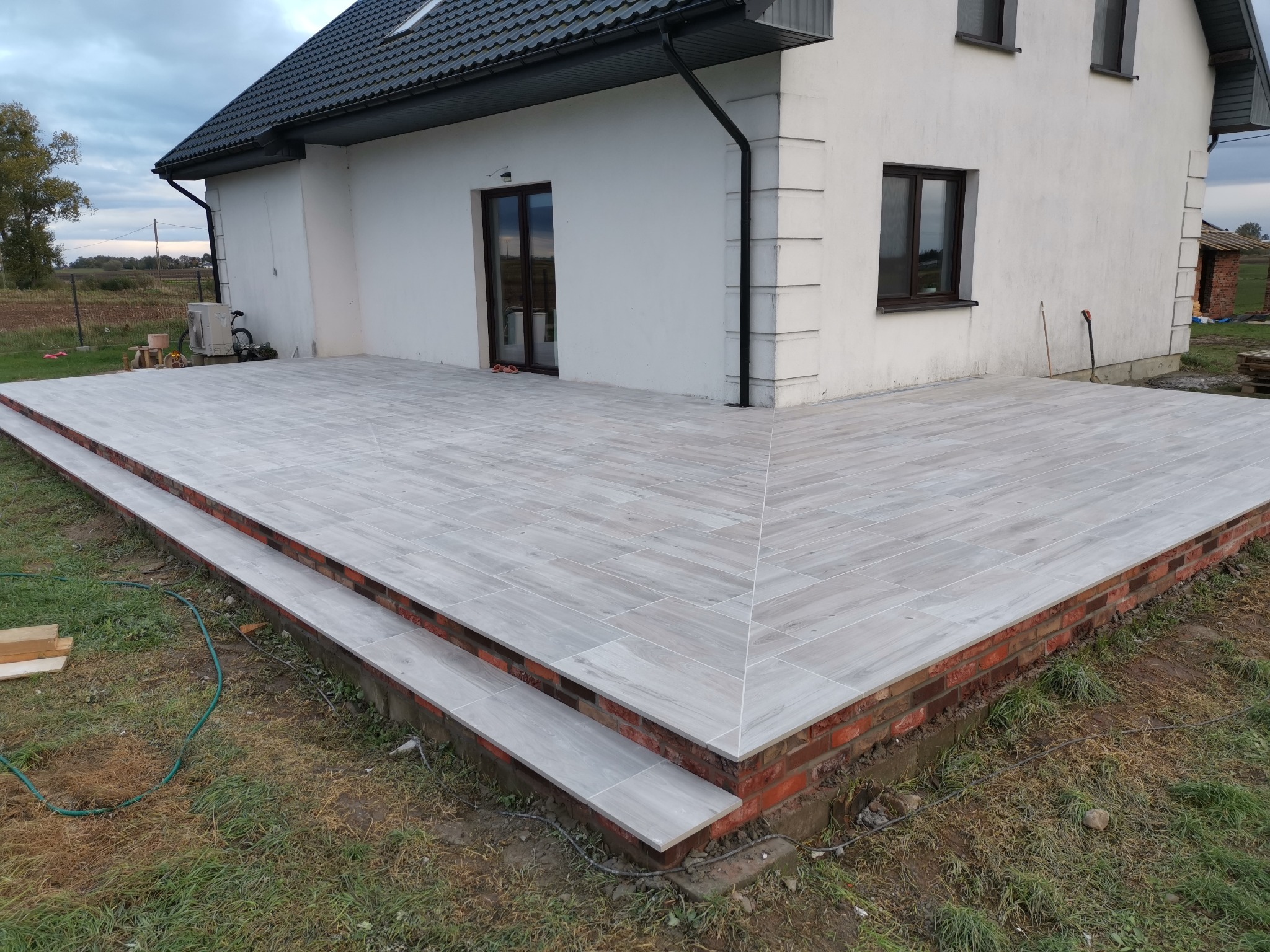 Nowoczesny taras z szarych płytek imitujących drewno, obrzeża z czerwonej cegły, widok na biały dom z ciemnymi oknami i dachem, trawnik w tle.