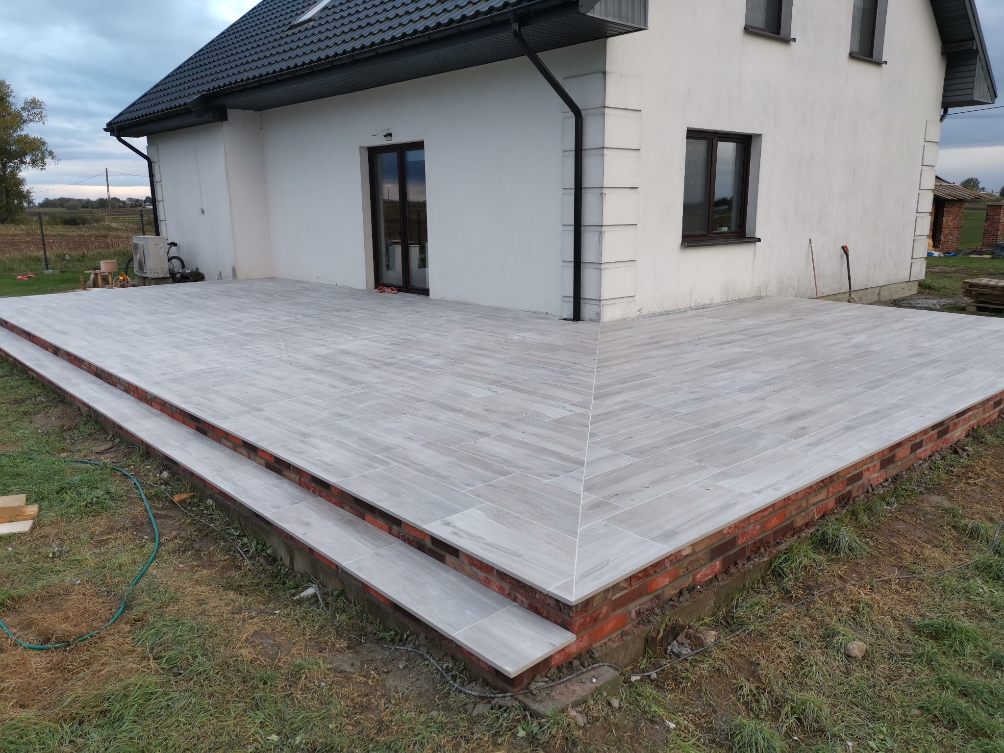 Wykończony taras z jasnoszarych płytek imitujących drewno, z dwustopniowymi schodami, obrzeżem z czerwonej cegły i widocznym fragmentem elewacji domu w tle.