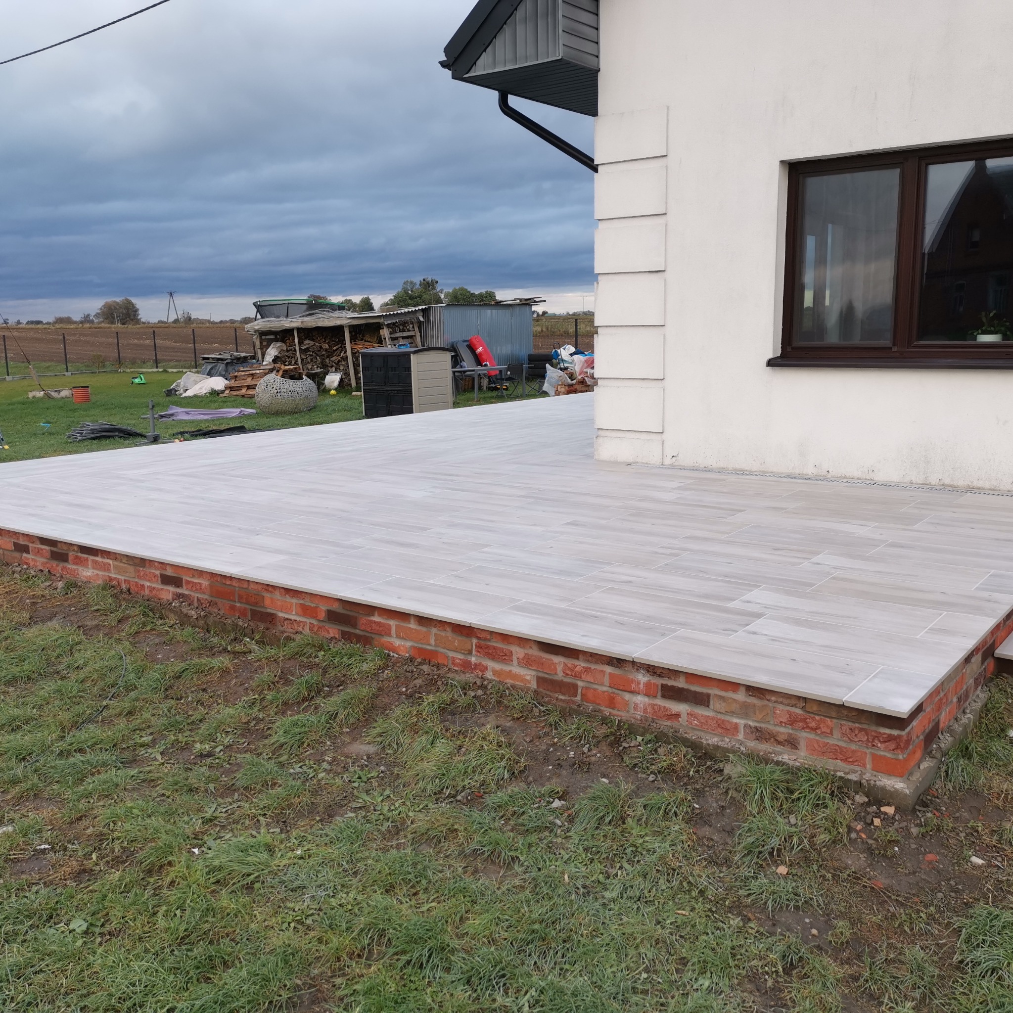 Wykończony taras z jasnych, podłużnych płytek imitujących drewno, obłożony czerwoną cegłą, przylegający do białego budynku z brązowym oknem i widokiem na podwórze z drewnem na opał pod pochmurnym...