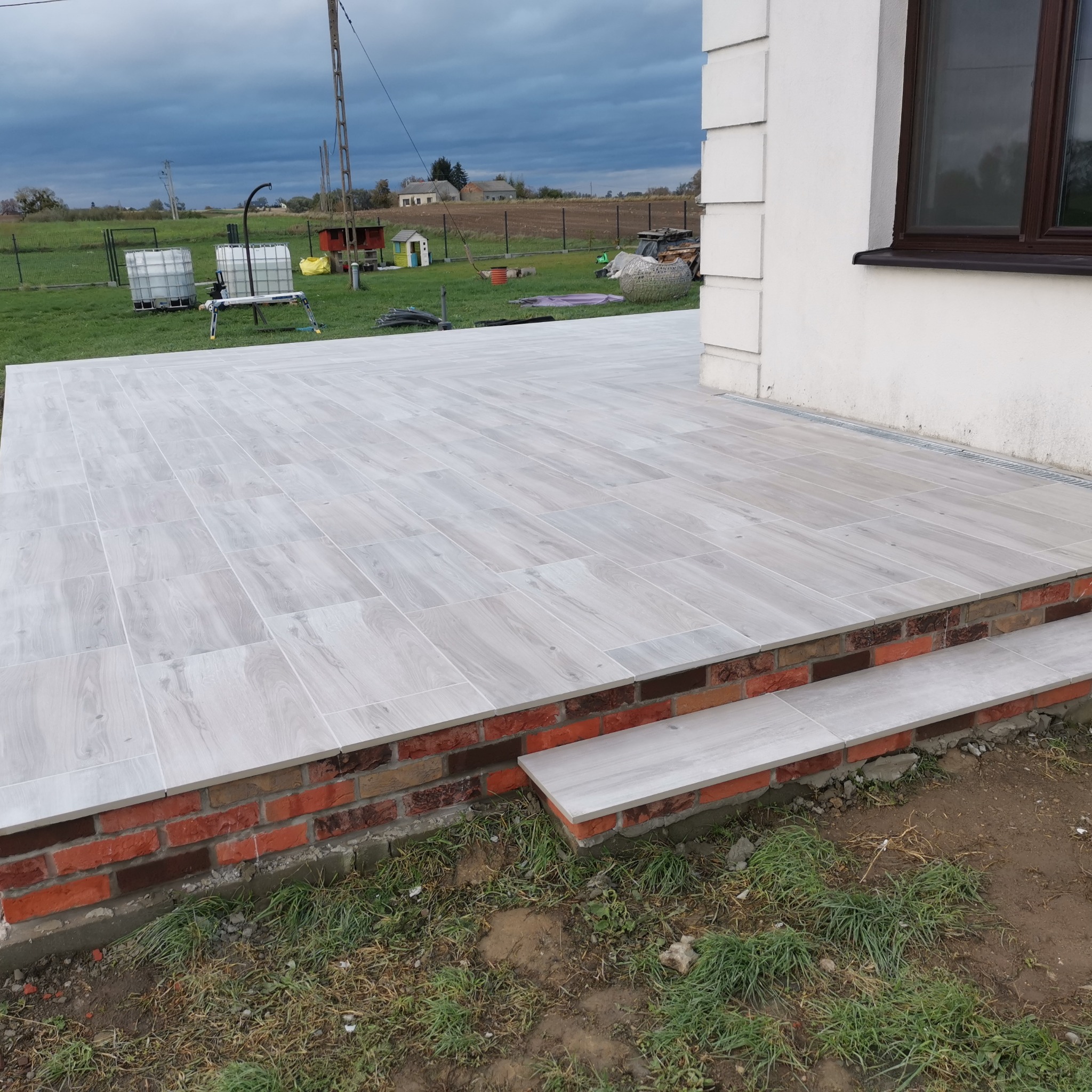 Wyłożony jasnoszarą, podłużną płytką taras z ceglanym obramowaniem i dwoma stopniami, z widokiem na trawnik i zabudowania gospodarcze w tle pod pochmurnym niebem.
