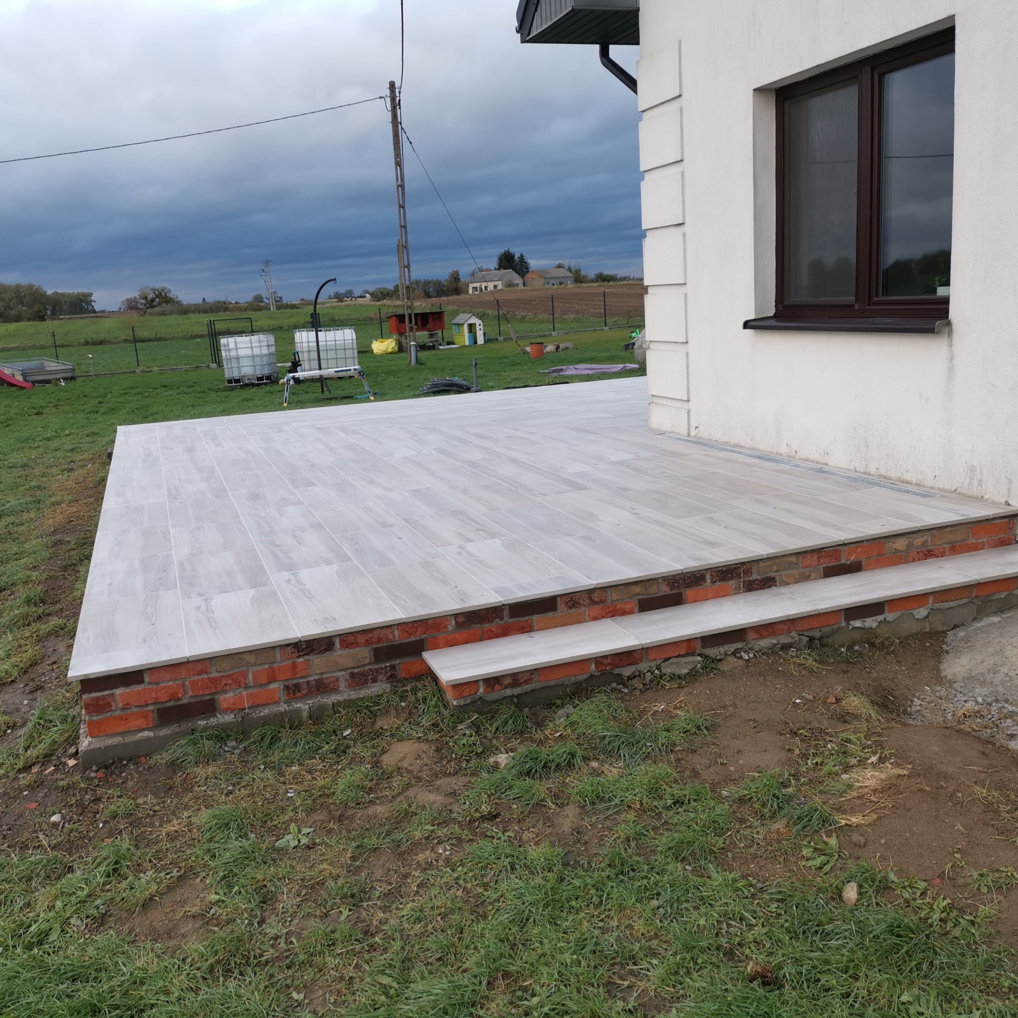 Wykończony taras z jasnoszarych płytek imitujących drewno, z dwoma stopniami obłożonymi tymi samymi płytkami, podmurówka z czerwonej cegły, widok na trawnik i zachmurzone niebo.