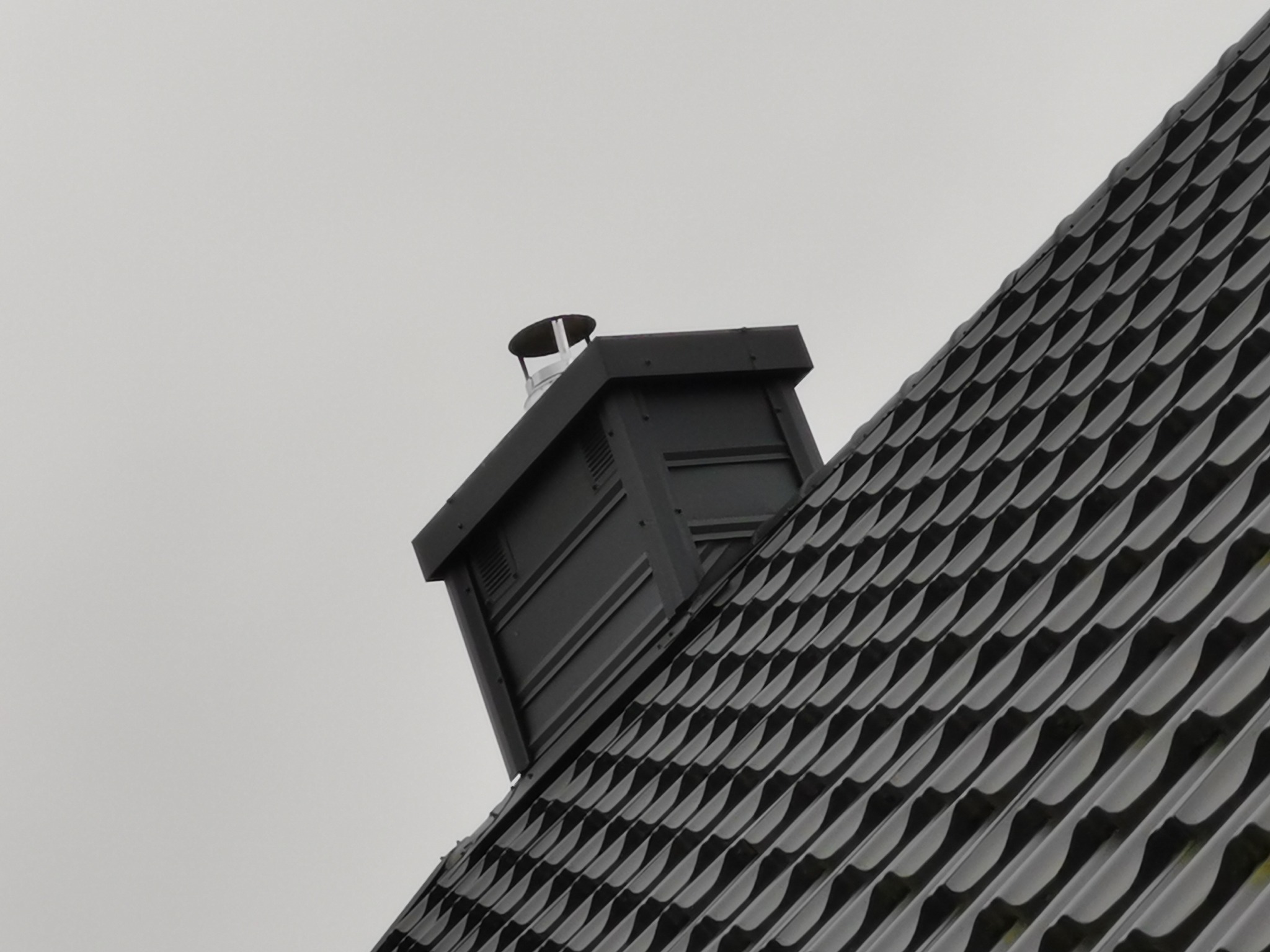 Grafitowy komin wentylacyjny z metalową nasadą na tle pochmurnego nieba i fragmentu dachu z blachy trapezowej w odcieniach szarości.