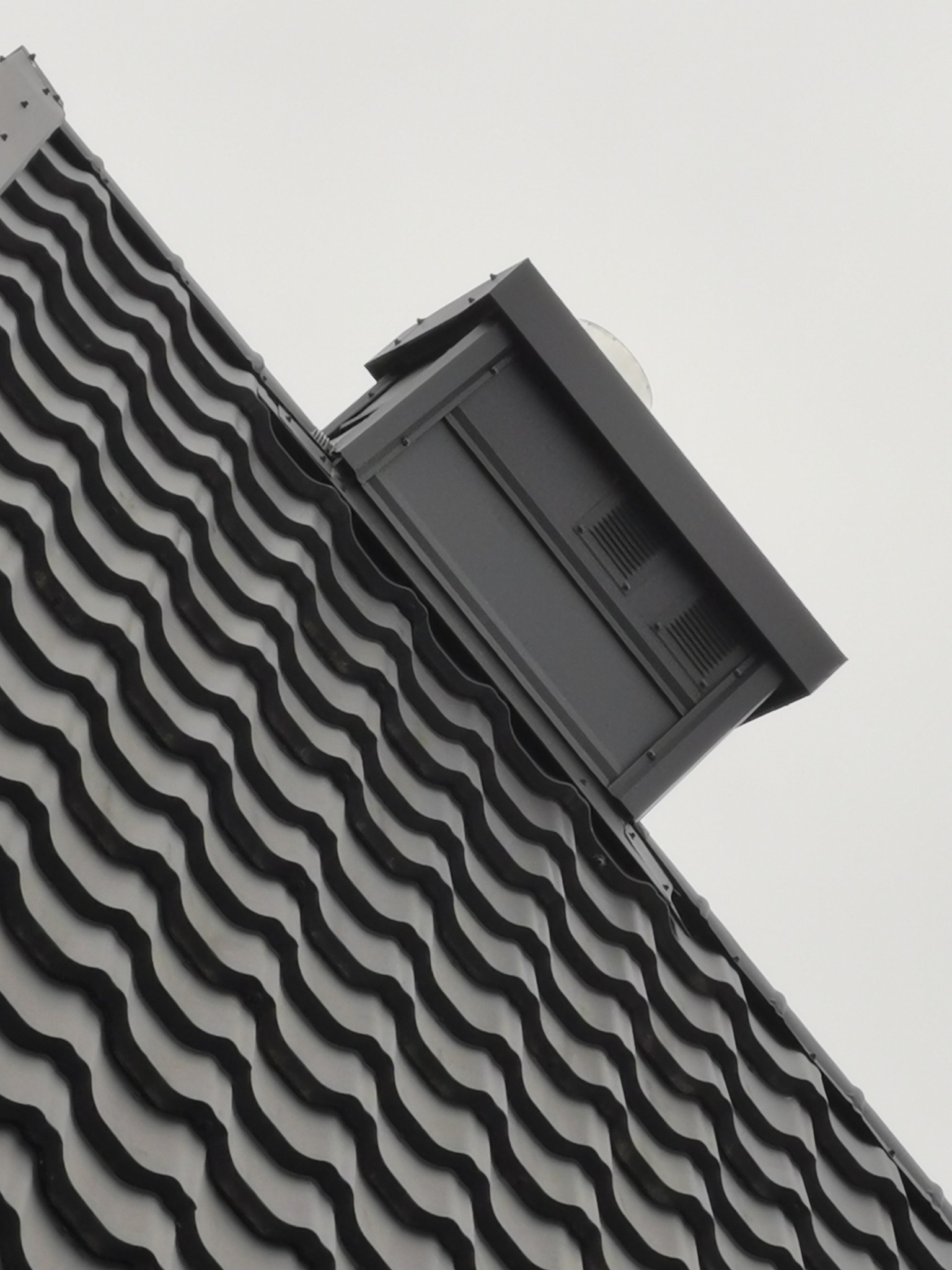 Fragment dachu z czarnej, falistej dachówki ceramicznej i czarnym kominkiem wentylacyjnym z widocznymi kratkami wentylacyjnymi, sfotografowany pod kątem.