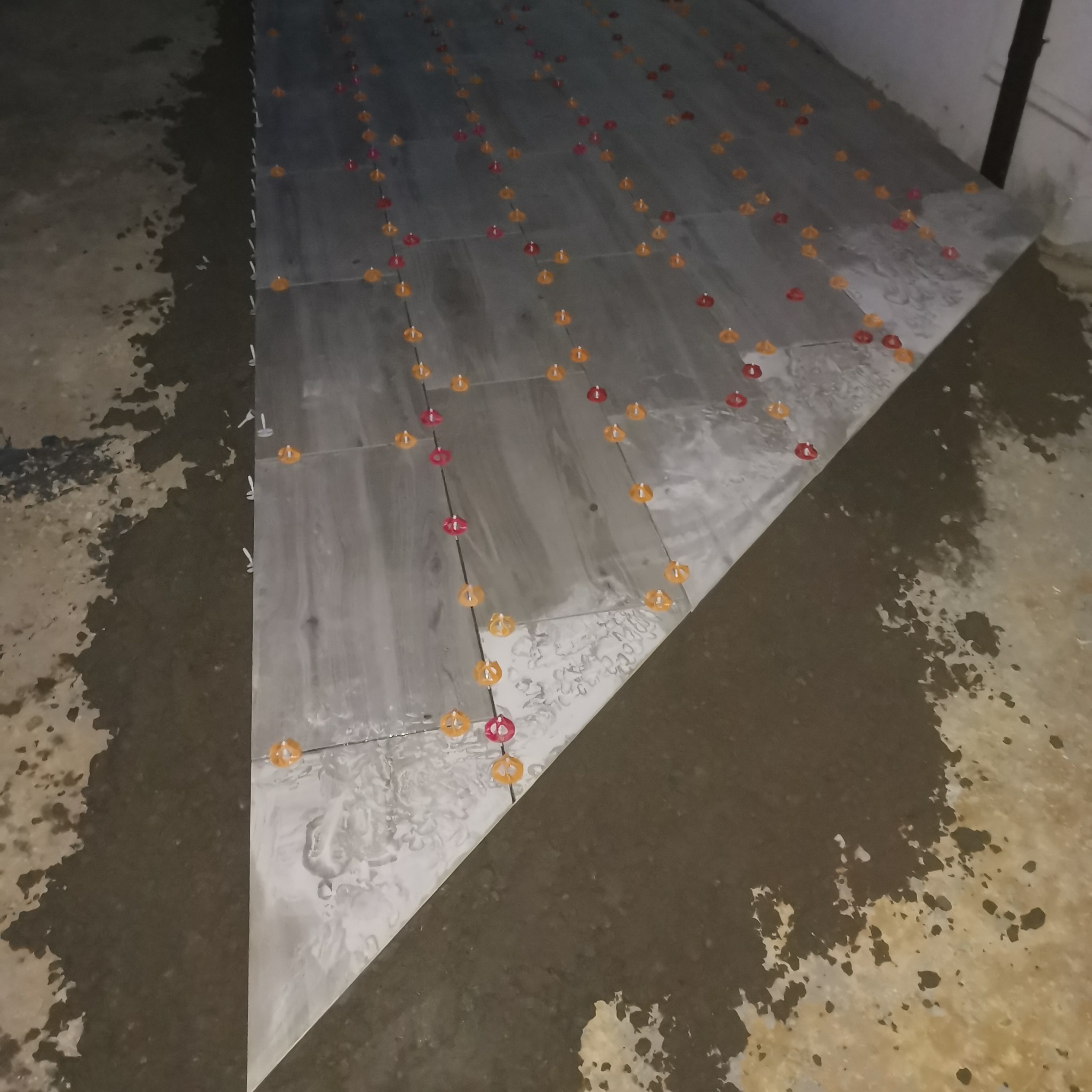 Układanie szarych płytek podłogowych z użyciem czerwonych i żółtych krzyżyków dystansowych oraz białych klinów poziomujących.