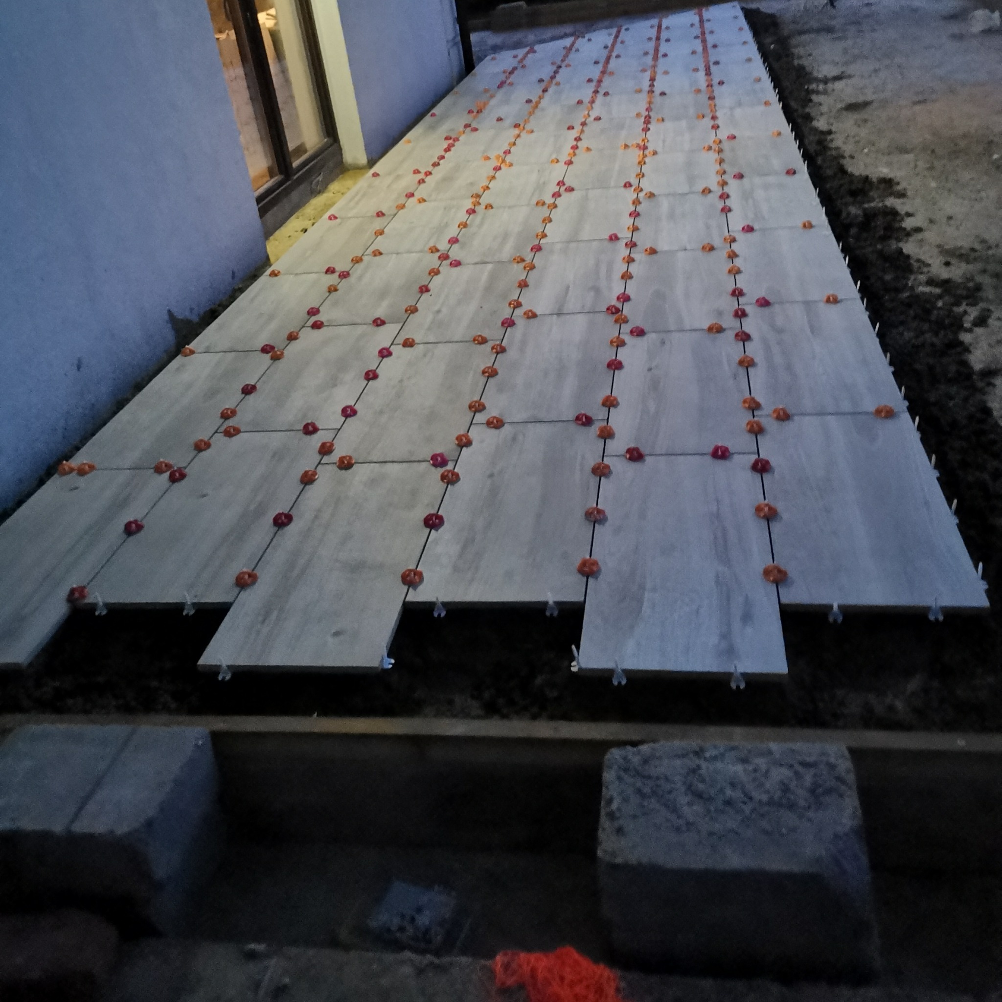 Układanie płytek podłogowych na zewnątrz budynku, z użyciem systemu poziomowania płytek (czerwone klipsy i czarne kliny), widoczne niedociągnięte płytki i przygotowane podłoże.