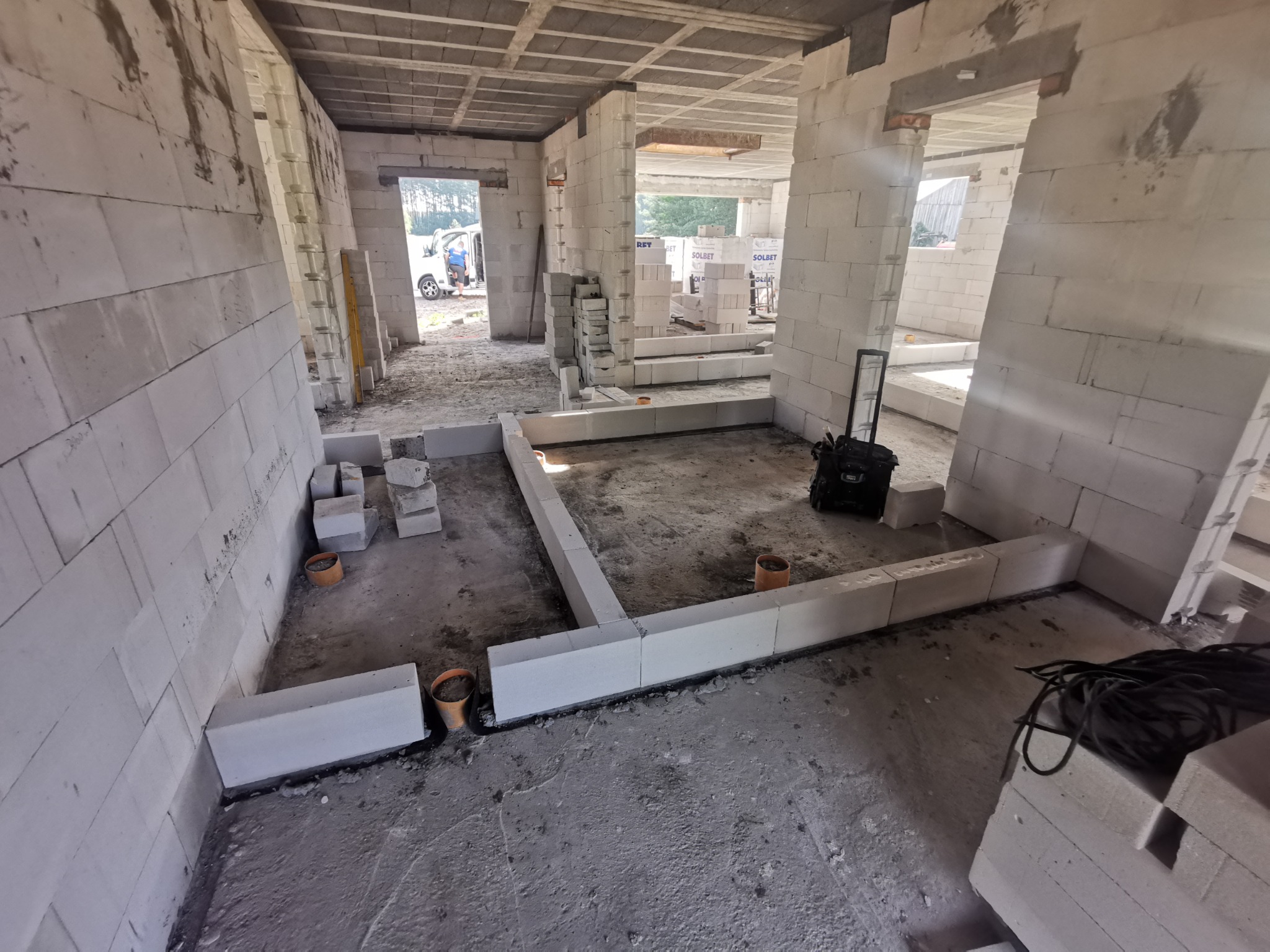 Wnętrze surowego domu w trakcie budowy z bloczków Solbet, widoczne fundamenty, ściany działowe i strop, na zewnątrz zaparkowany biały bus.