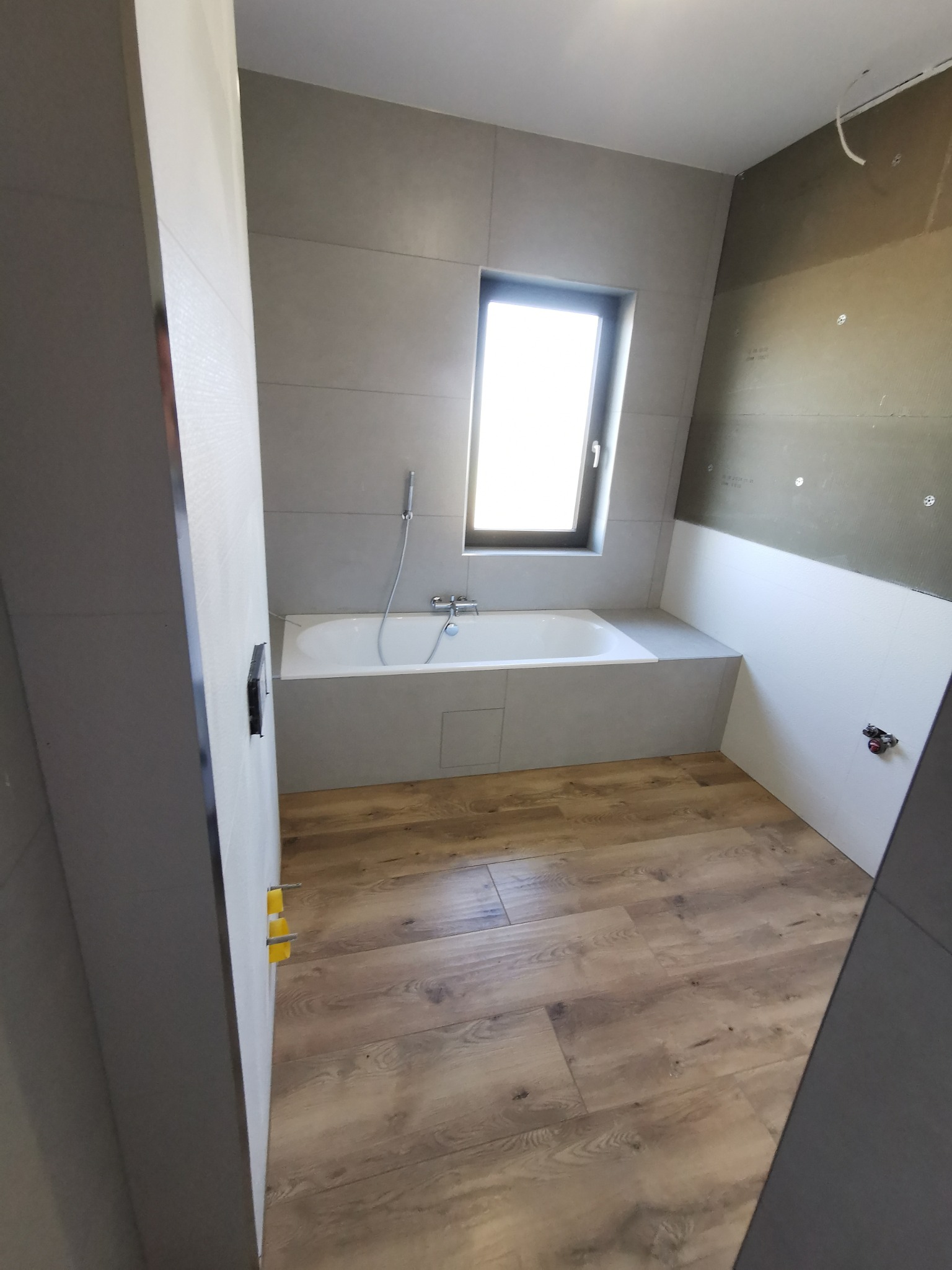 Wnętrze łazienki w trakcie wykańczania w Obrowie: widoczna wanna pod oknem, szare płytki na ścianach, drewnopodobna podłoga i fragment ściany z izolacją.