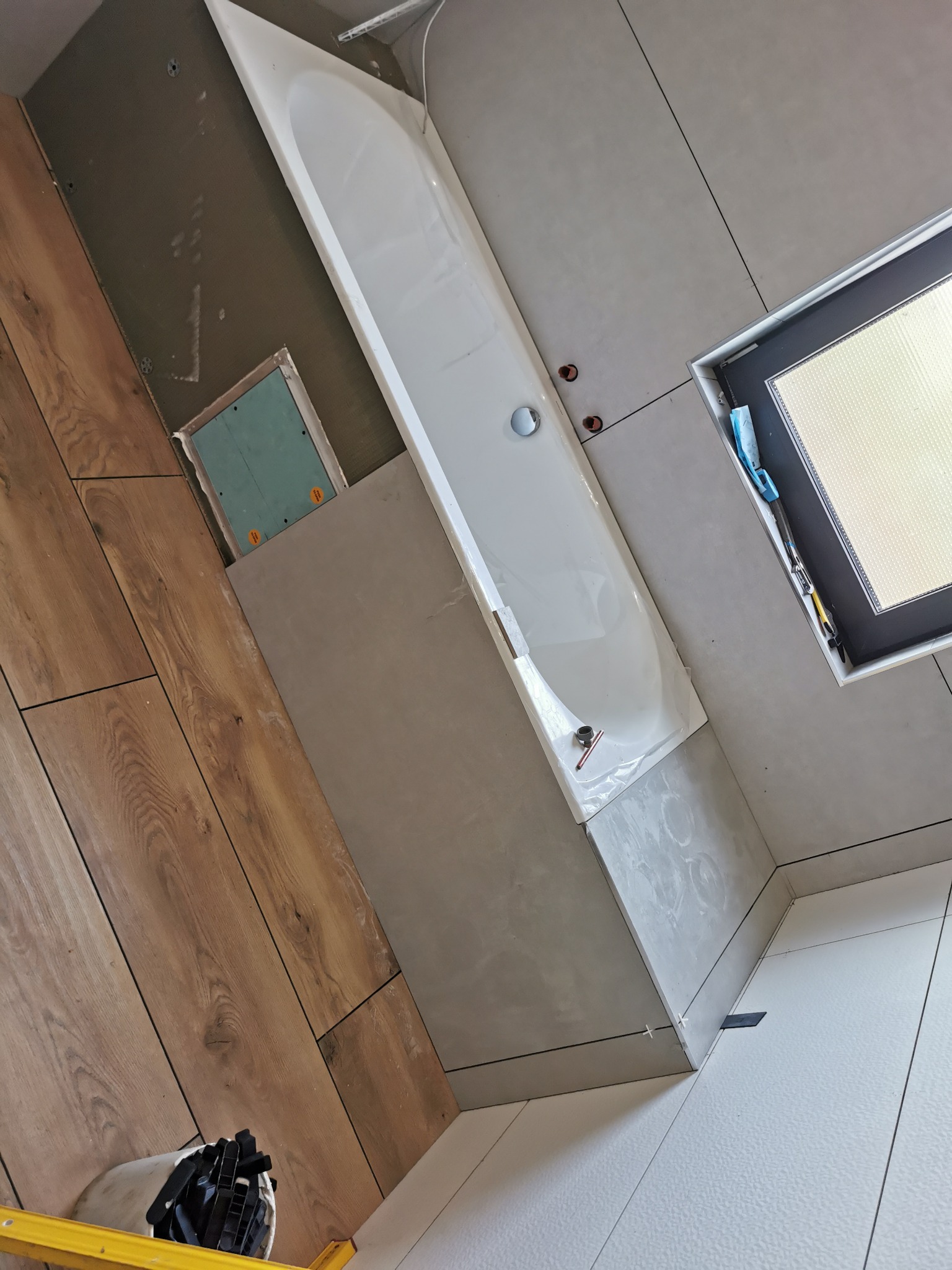 Montaż wanny w łazience, widoczne płytki imitujące drewno na ścianie, szare płytki ścienne i białe podłogowe, okno z zamontowaną roletą, poziomica i akcesoria glazurnicze.