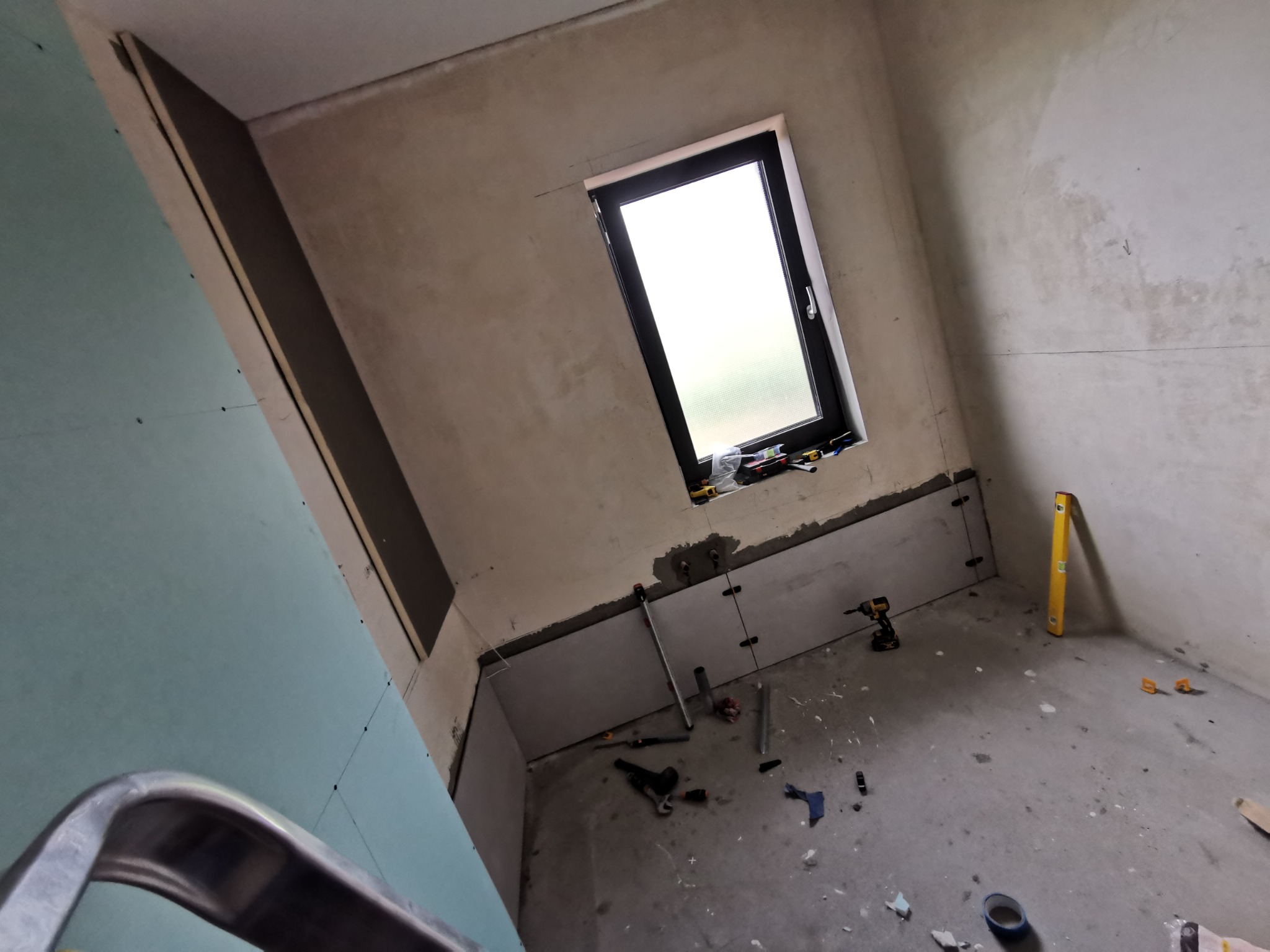 Wnętrze łazienki w trakcie prac wykończeniowych: widoczne płyty gipsowo-kartonowe na ścianach, okno, narzędzia budowlane na podłodze, poziomica przy ścianie, oraz fragment drabiny w lewym dolnym rogu.