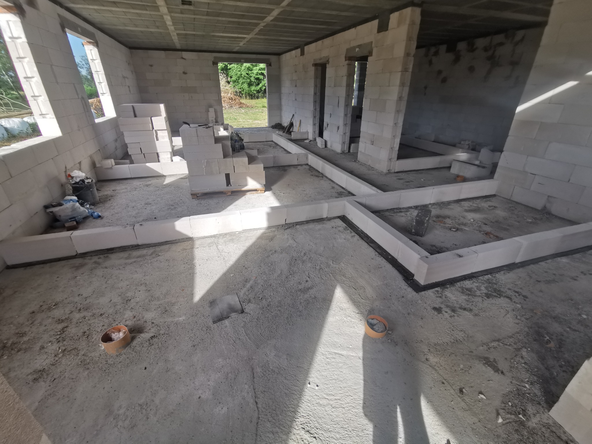 Surowy stan wnętrza domu w trakcie budowy, ściany z białych bloczków, widoczne fundamenty i otwory okienne, betonowa podłoga z elementami instalacji, częściowo ułożone bloczki na ścianach.