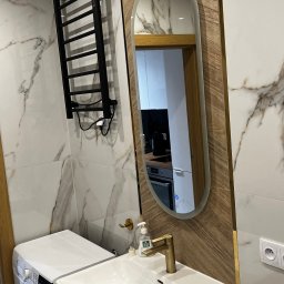 Marcin Szalanski - Nowoczesna łazienka z pralką obok szafki z umywalką, czarny grzejnik drabinkowy na ścianie z marmurowym wzorem, owalne lustro z drewnianą ramą odbijające wnętrze kuchni.