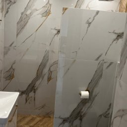 Marcin Szalanski - Nowoczesna łazienka z marmurowymi płytkami na ścianach i podłodze, złota armatura prysznicowa, biała umywalka z szafką i toaleta, podłoga z drewnopodobnych płytek ułożonych w jodełkę w strefie...