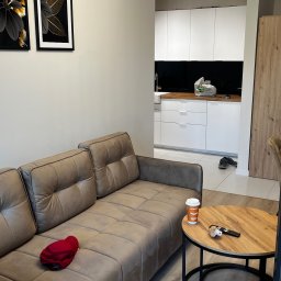 Marcin Szalanski - Jasny salon z szarobrązową sofą, okrągłym stolikiem kawowym z pilotem i kubkiem, oraz widokiem na białą kuchnię z czarnym blatem i drewnianą ścianą, oświetlony nowoczesną lampą sufitową.