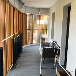 Marcin Szalanski - Zadaszony balkon z drewnianą ażurową balustradą, szarą podłogą z desek kompozytowych i widoczną suszarką na pranie oraz kartonami z akcesoriami do sprzątania.