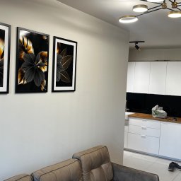 Marcin Szalanski - Salon z beżową sofą i trzema obrazami z motywem kwiatowym w czarnych ramach na ścianie, w tle widoczna biała kuchnia z czarnym panelem między szafkami.