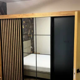 Nowoczesna szafa wnękowa z przesuwnymi drzwiami, częściowo lustrzanymi, z drewnianymi akcentami, na tle ściany z dekoracyjnych paneli 3D w odcieniach czerni i złota.