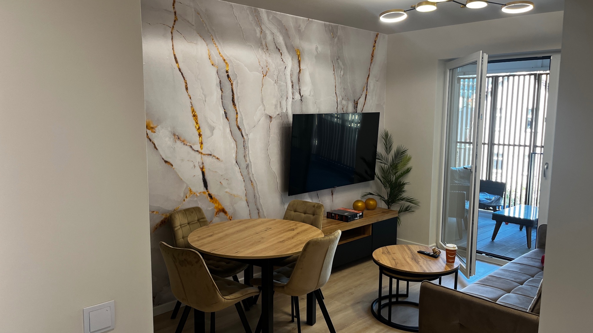 Nowoczesne wnętrze pokoju dziennego z okrągłym stołem i krzesłami na tle ściany z marmurowym wzorem i telewizorem, widok na balkon przez otwarte drzwi.