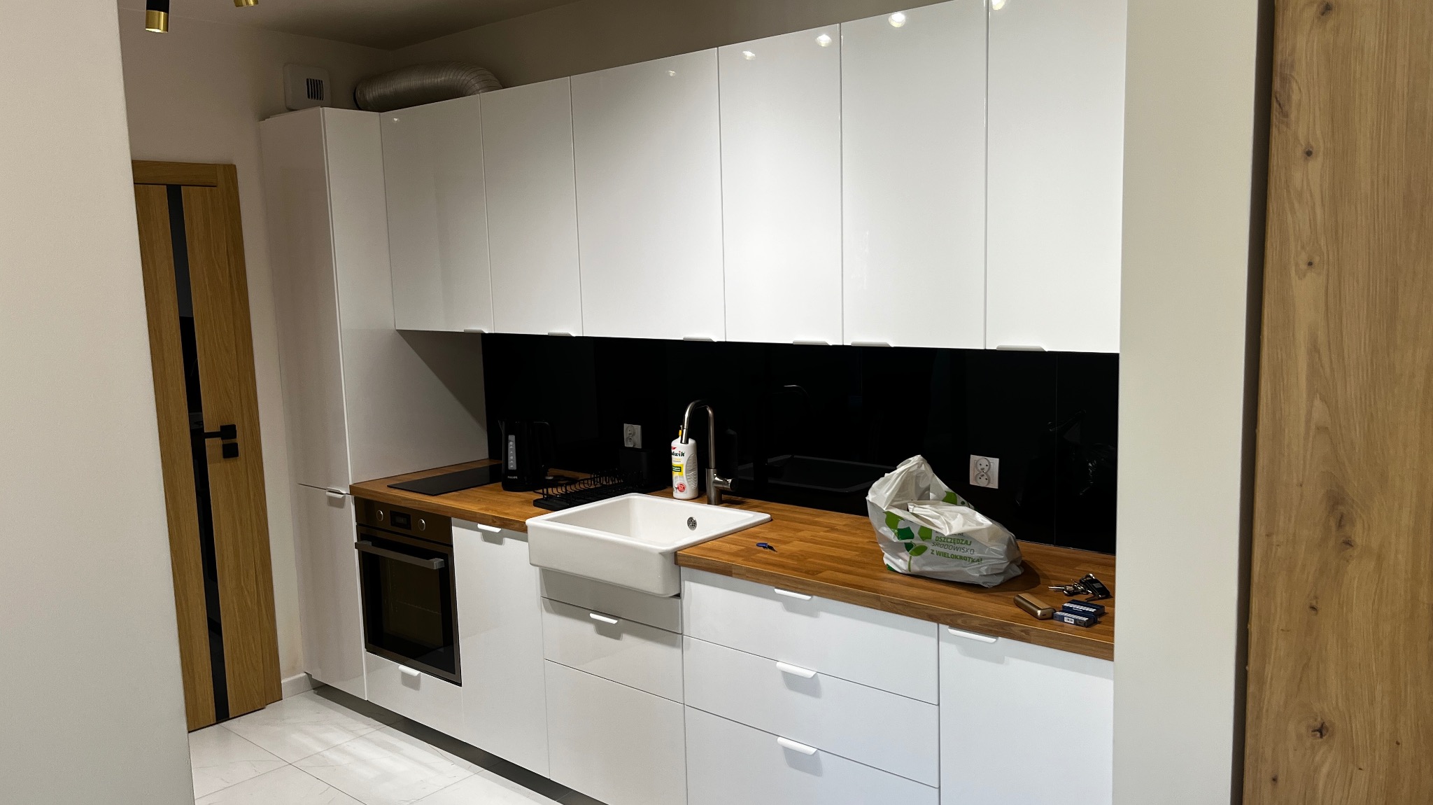 Nowoczesna, biała kuchnia z drewnianym blatem, ceramicznym zlewem typu farmhouse, czarnym panelem nad blatem i widoczną torbą zakupową na blacie.