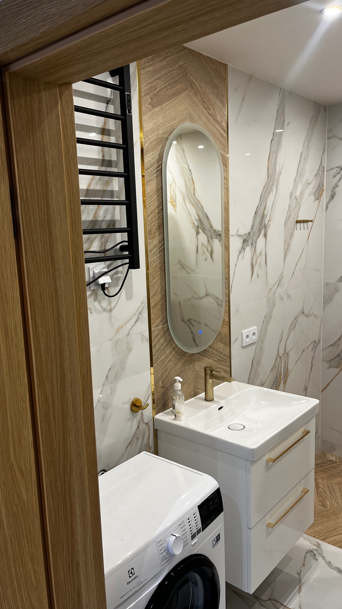 Nowoczesna łazienka z marmurowymi płytkami, owalnym lustrem z podświetleniem LED, białą umywalką z szafką i pralką Electrolux, widoczna przez drewniane futryny drzwi.