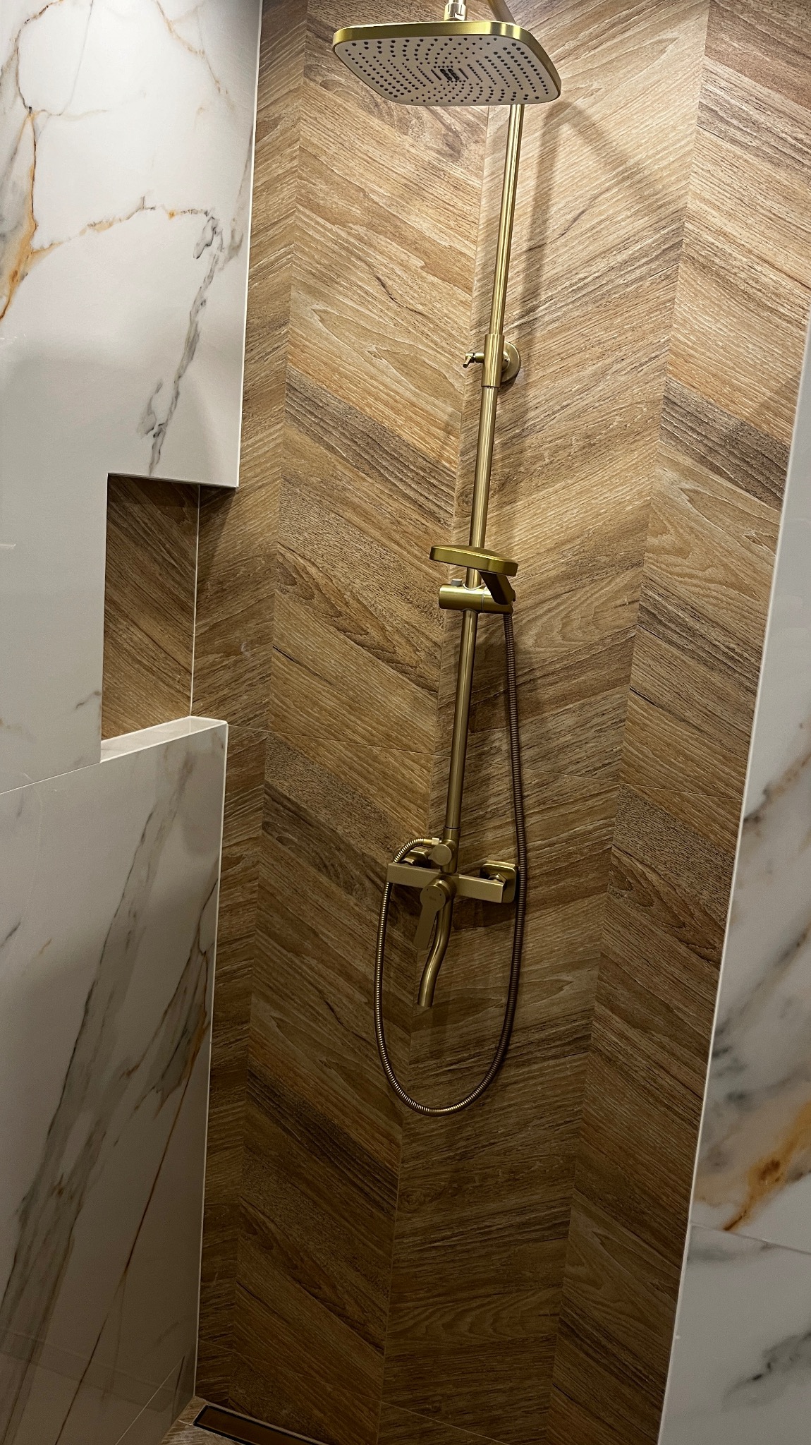 Złota bateria prysznicowa z deszczownicą na ścianie wyłożonej płytkami imitującymi drewno w jodełkę, obok ściana z białego marmuru z wnęką.