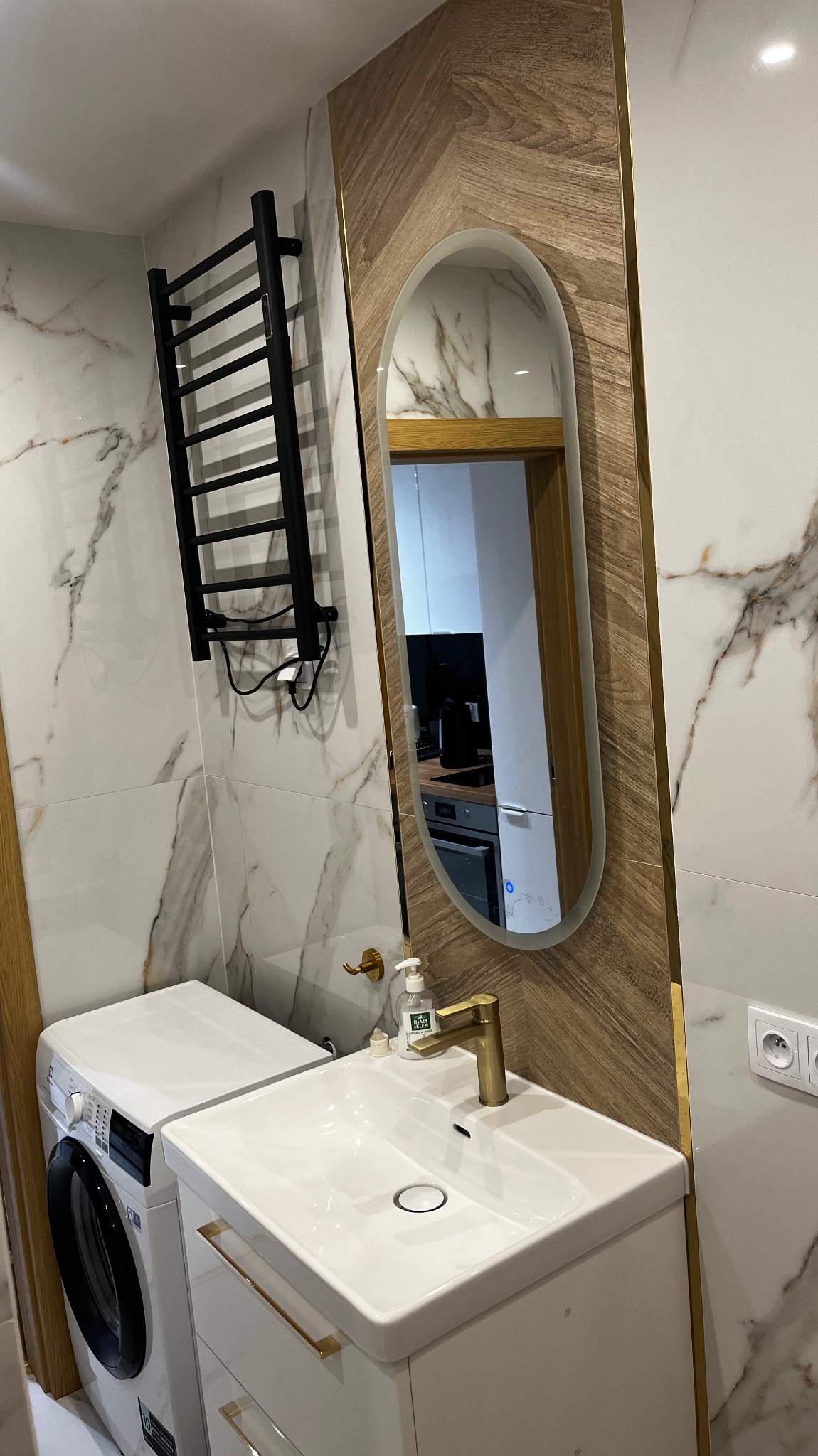 Nowoczesna łazienka z pralką obok szafki z umywalką, czarny grzejnik drabinkowy na ścianie z marmurowym wzorem, owalne lustro z drewnianą ramą odbijające wnętrze kuchni.