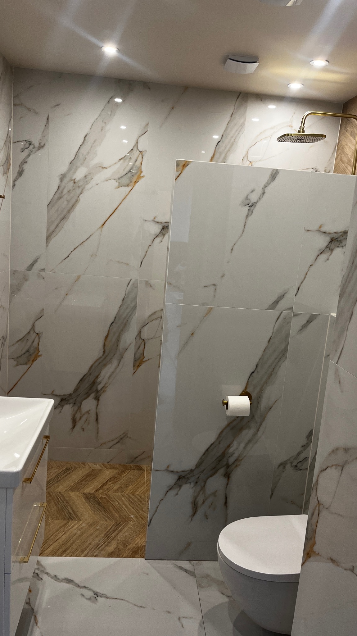 Nowoczesna łazienka z marmurowymi płytkami na ścianach i podłodze, złota armatura prysznicowa, biała umywalka z szafką i toaleta, podłoga z drewnopodobnych płytek ułożonych w jodełkę w strefie...