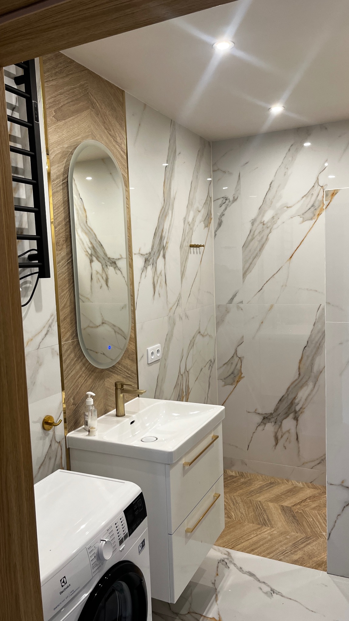Nowoczesna łazienka z marmurowymi płytkami, podświetlanym lustrem w złotej ramie, białą szafką umywalkową z złotymi uchwytami i pralką Electrolux.