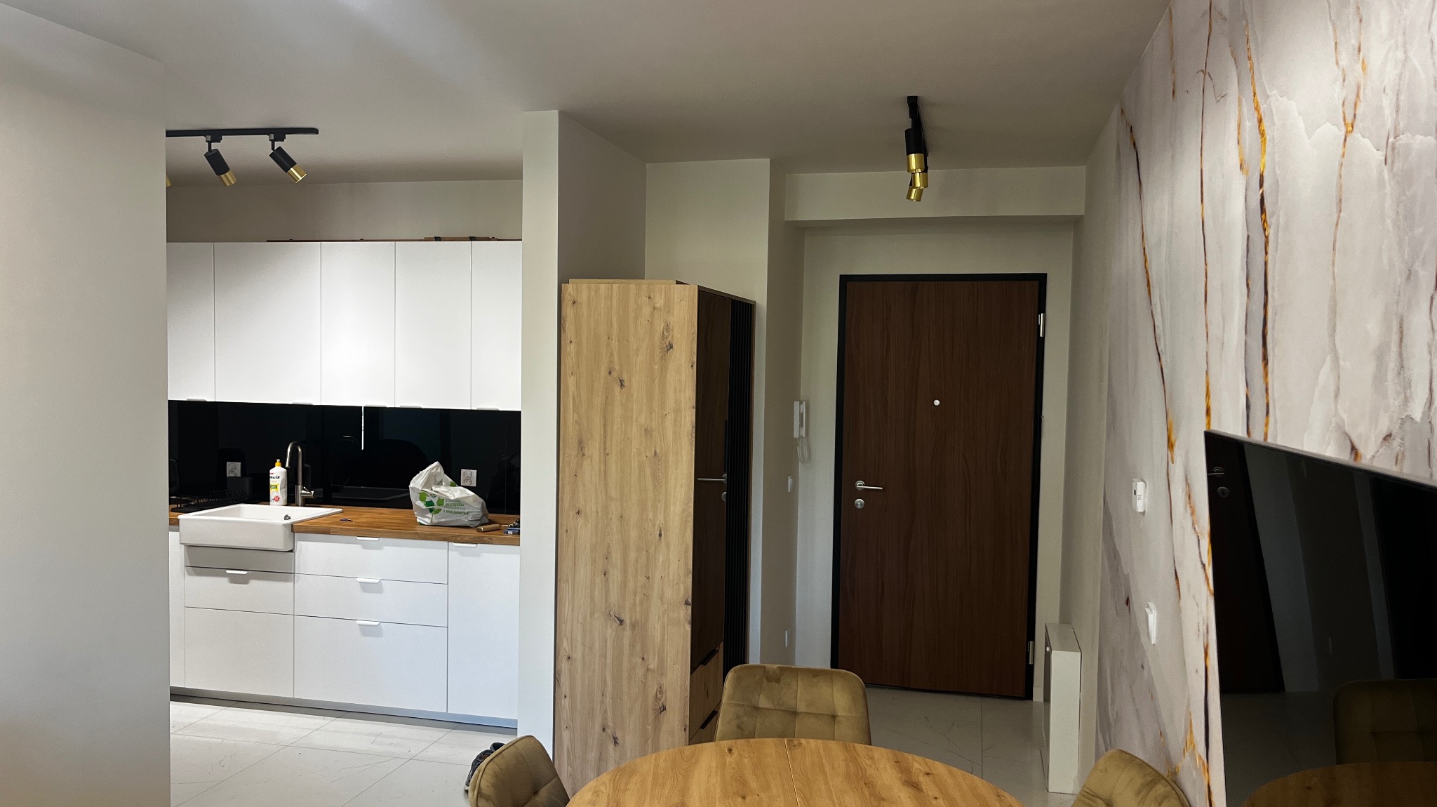 Nowoczesne wnętrze: biała kuchnia z czarnym panelem, drewniany blat, okrągły stół z krzesłami, drzwi wejściowe i ściana z marmurowym wzorem.