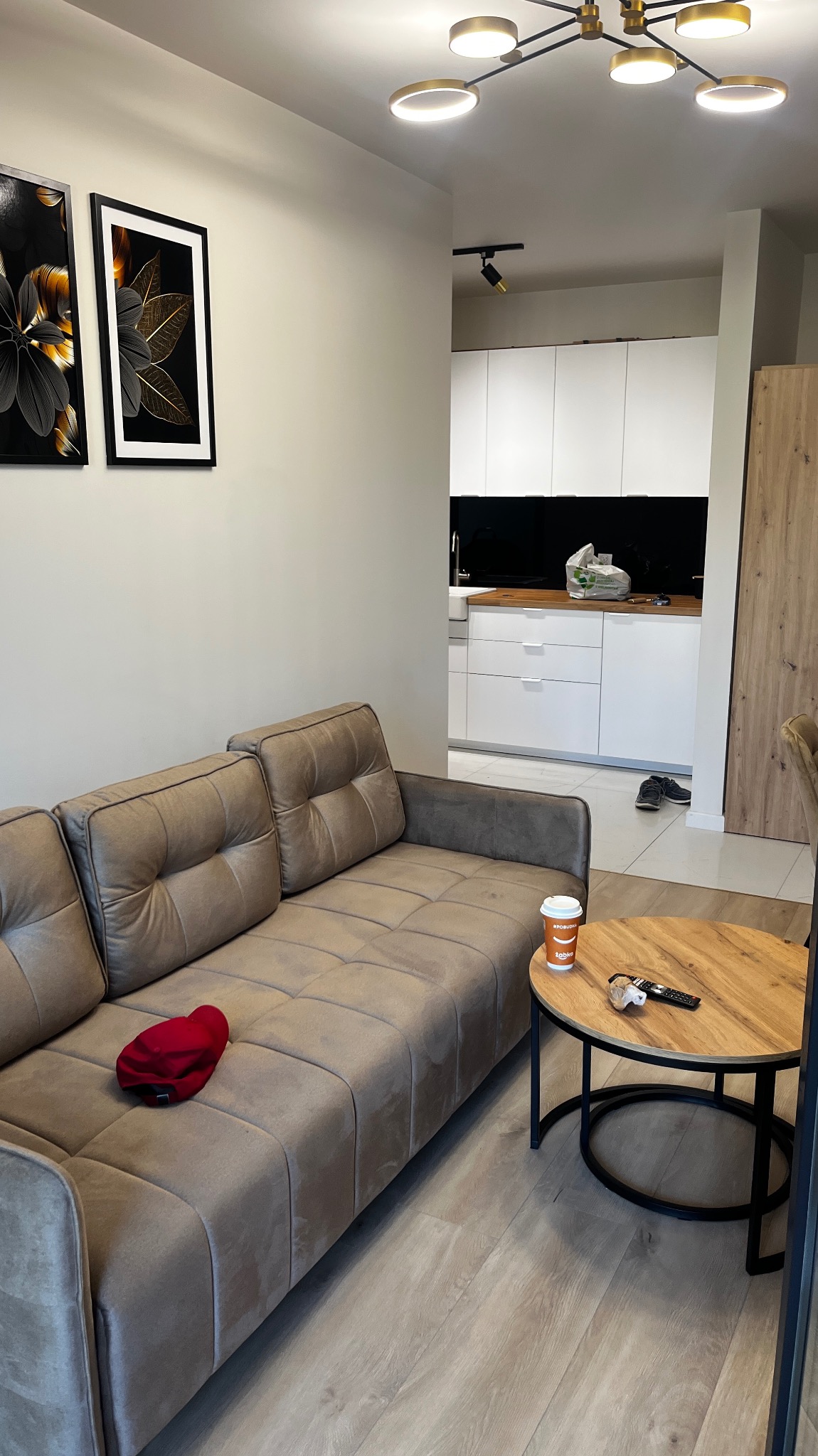 Jasny salon z szarobrązową sofą, okrągłym stolikiem kawowym z pilotem i kubkiem, oraz widokiem na białą kuchnię z czarnym blatem i drewnianą ścianą, oświetlony nowoczesną lampą sufitową.