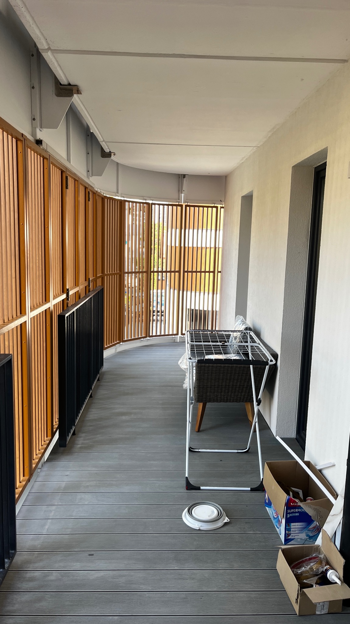 Zadaszony balkon z drewnianą ażurową balustradą, szarą podłogą z desek kompozytowych i widoczną suszarką na pranie oraz kartonami z akcesoriami do sprzątania.