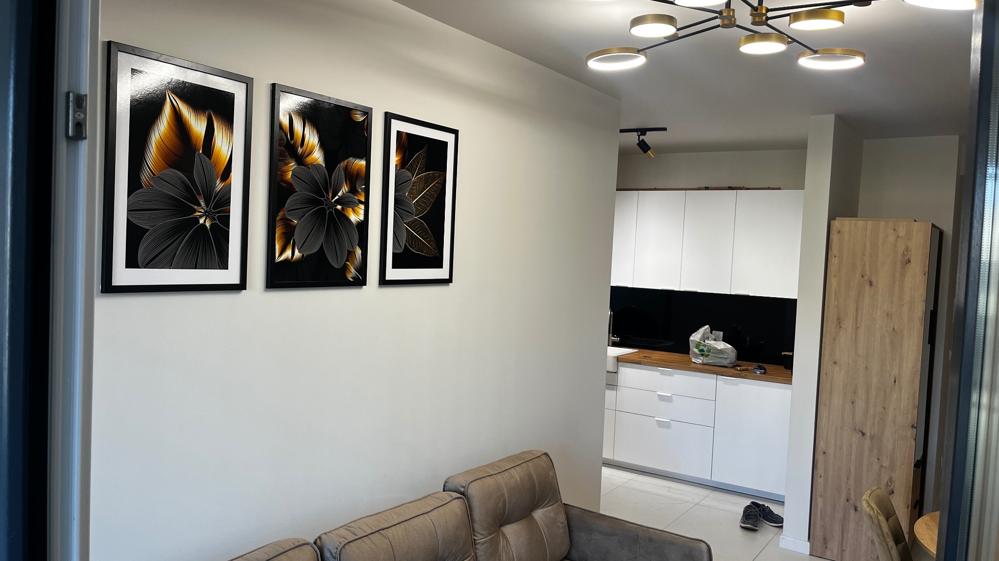 Salon z beżową sofą i trzema obrazami z motywem kwiatowym w czarnych ramach na ścianie, w tle widoczna biała kuchnia z czarnym panelem między szafkami.