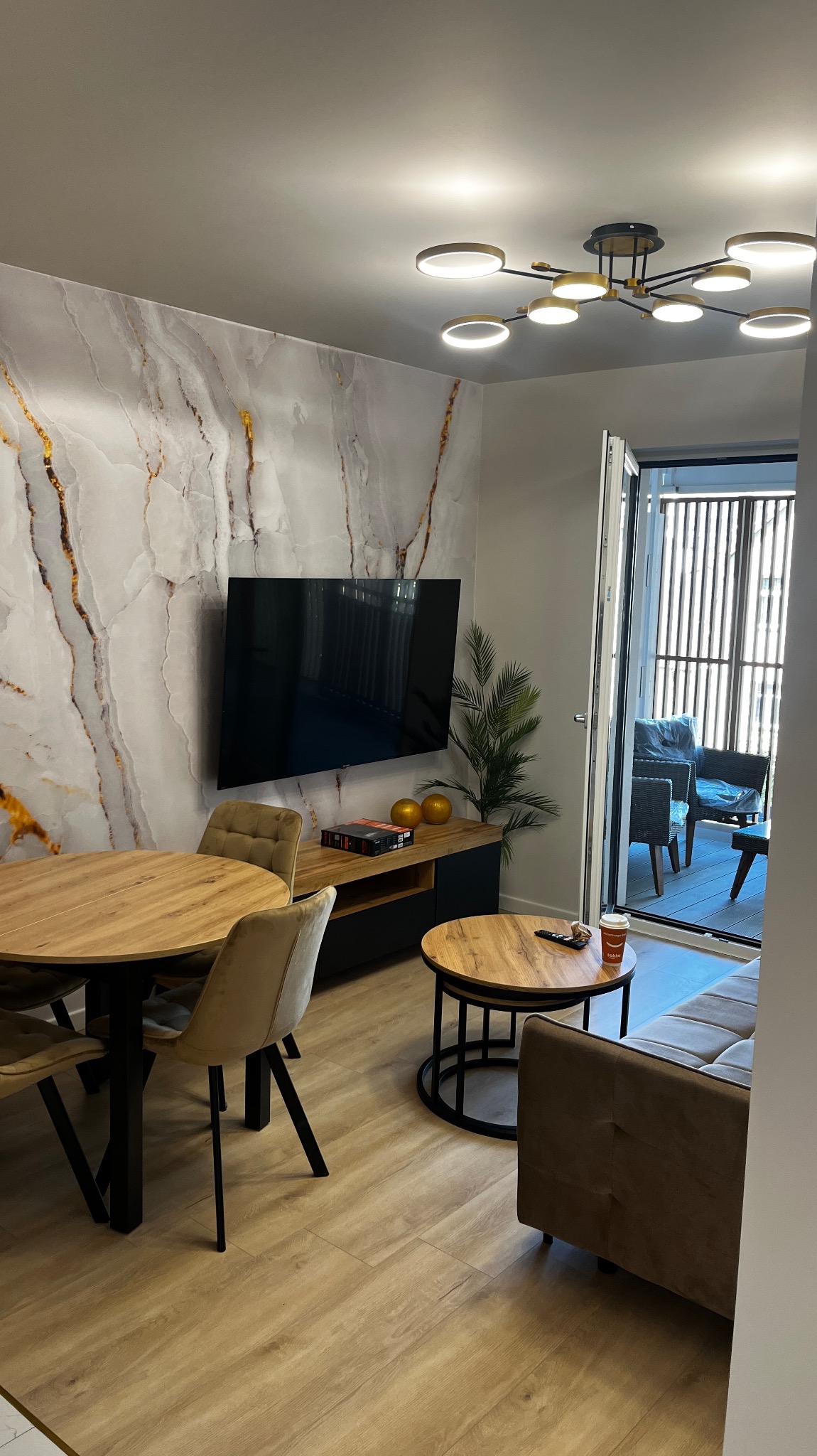 Nowoczesne wnętrze salonu z okrągłym stołem, telewizorem na ścianie z marmurowym wzorem i wyjściem na balkon z meblami wypoczynkowymi.