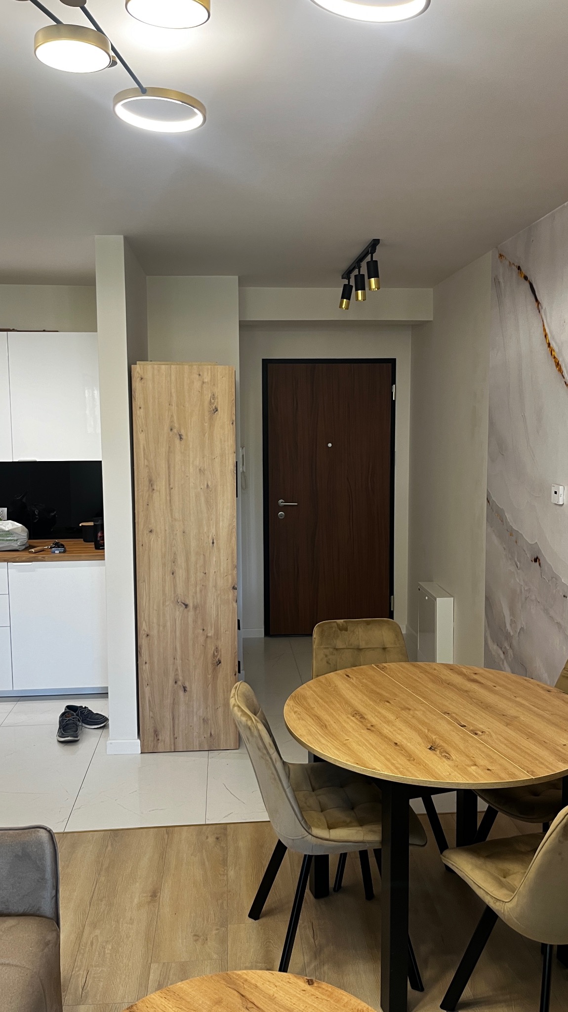 Jasne wnętrze mieszkania z okrągłym drewnianym stołem, welurowe krzesła, panele podłogowe w odcieniach drewna, drzwi wewnętrzne z ciemnego drewna oraz dekoracyjna tapeta na ścianie z motywem...