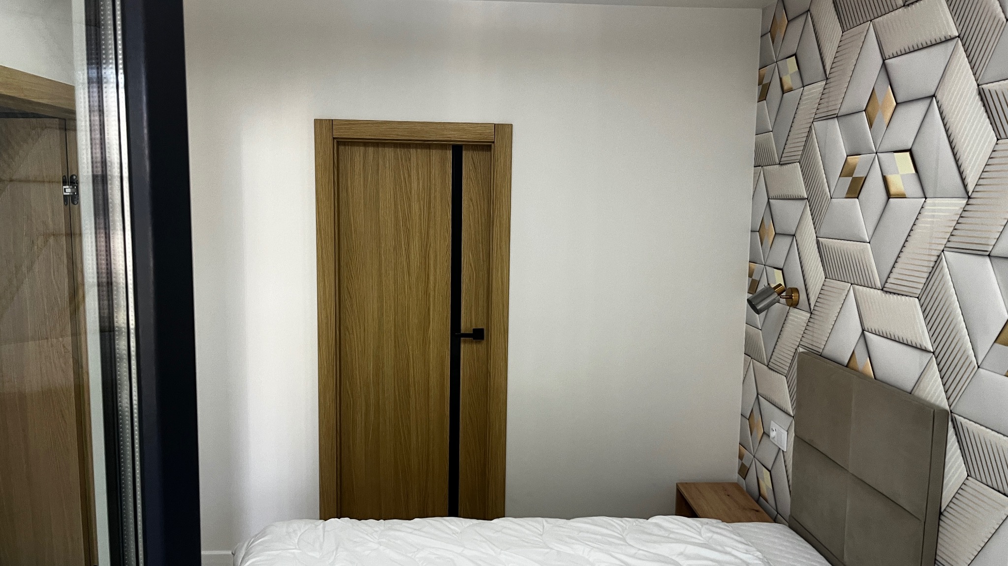 Zamknięte drzwi wewnętrzne z drewnianą okleiną i czarnym akcentem, widoczne na tle białej ściany i dekoracyjnej ściany z geometrycznym wzorem 3D w odcieniach bieli i złota, z fragmentem łóżka...