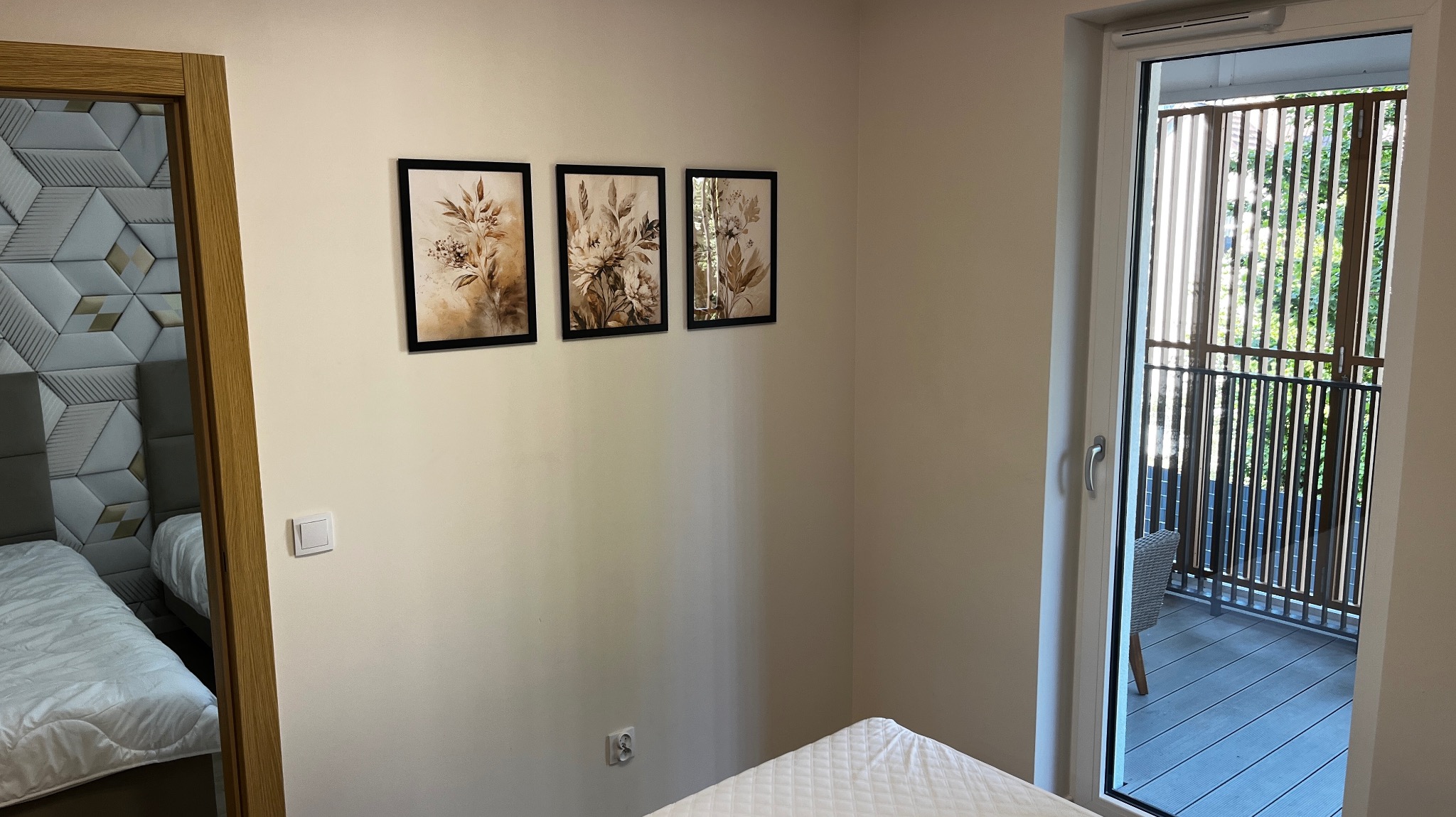 Nowoczesna sypialnia z wyjściem na balkon, widoczne trzy obrazy z motywami roślinnymi na ścianie i dekoracyjna ściana za łóżkiem z geometrycznym wzorem.