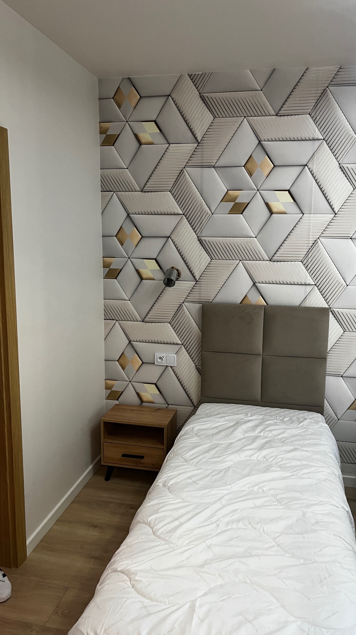 Nowoczesna sypialnia z dekoracyjną ścianą z paneli 3D w geometryczne wzory z akcentami złota, drewnianą szafką nocną i łóżkiem z pikowaną białą kołdrą.