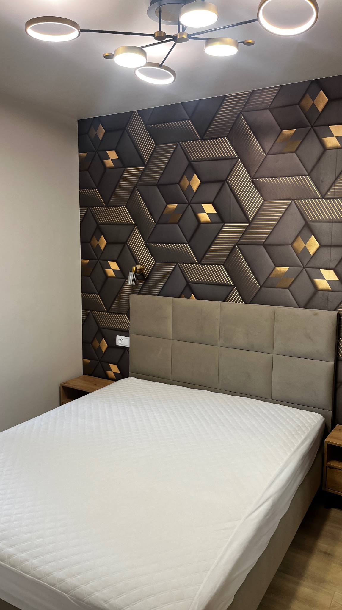 Sypialnia z łóżkiem i dekoracyjną ścianą z paneli 3D w geometryczne wzory w odcieniach brązu i złota, oświetlona nowoczesną lampą sufitową