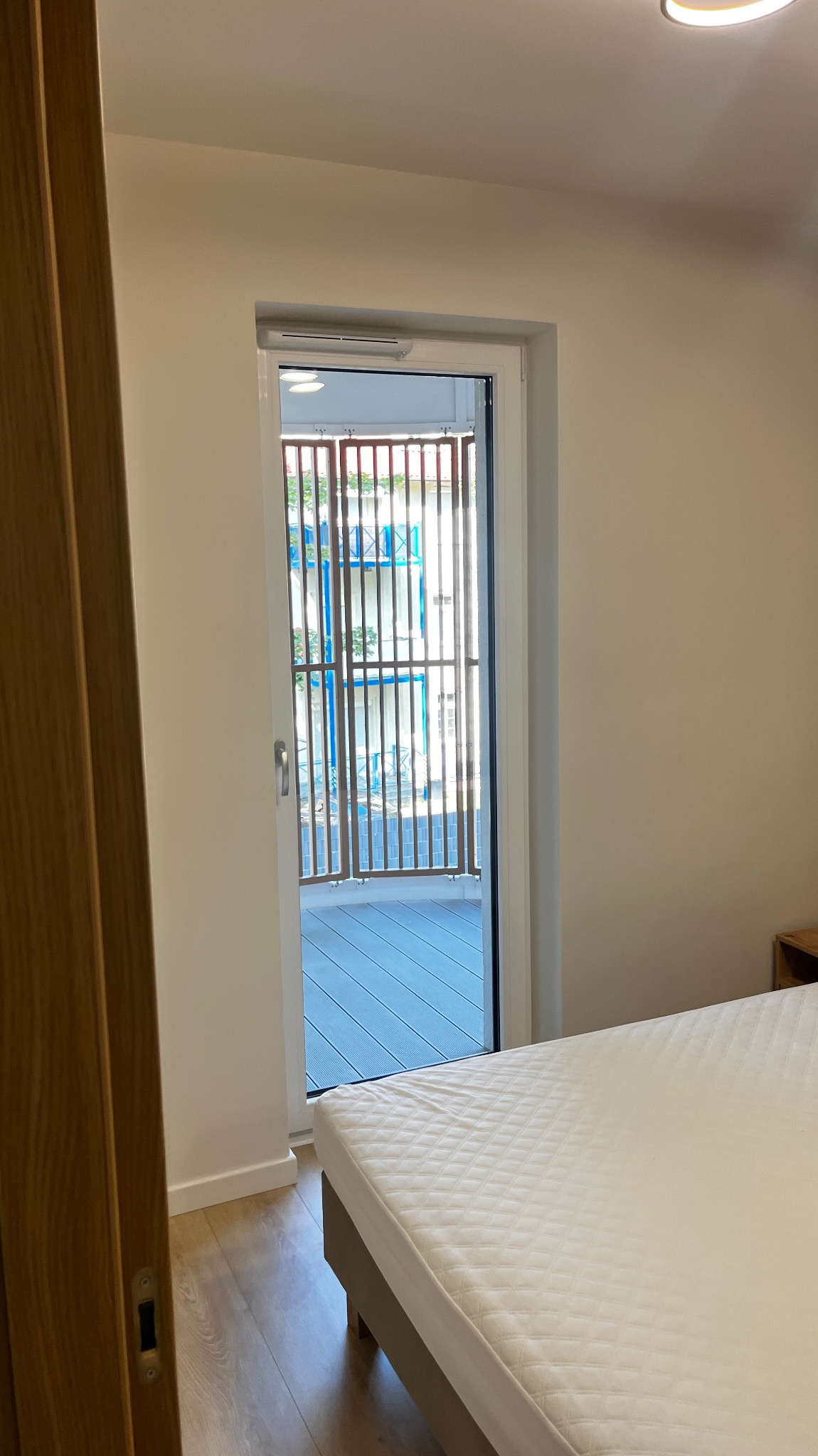 Wnętrze sypialni z widokiem na balkon z drewnianą balustradą i niebieską podłogą, przez otwarte drzwi balkonowe, widoczny fragment łóżka z białą pościelą.