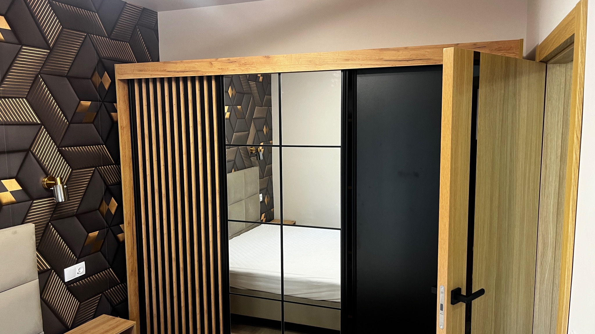 Nowoczesna szafa wnękowa z przesuwnymi drzwiami, częściowo lustrzanymi, z drewnianymi akcentami, na tle ściany z dekoracyjnych paneli 3D w odcieniach czerni i złota.