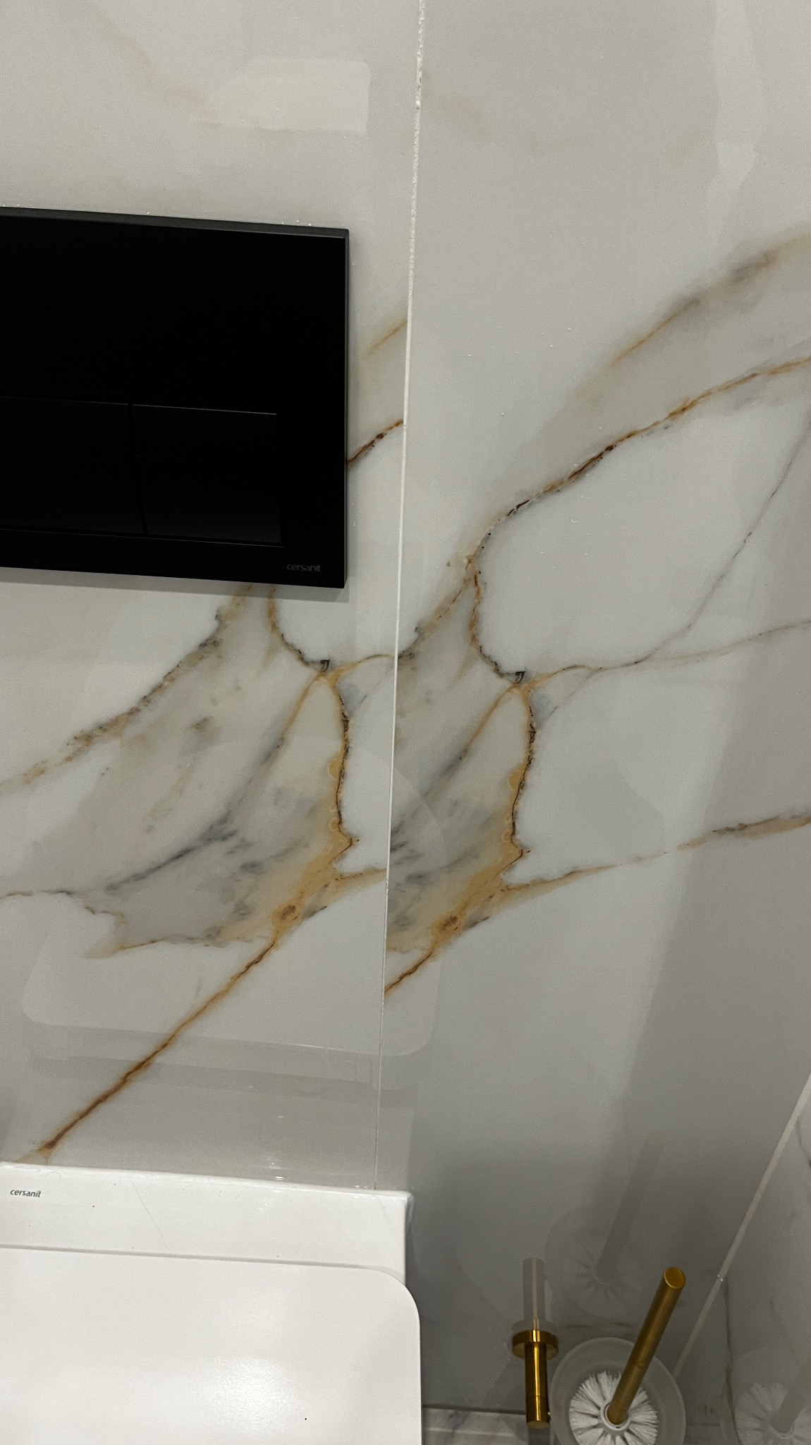 Fragment łazienki z widocznym białym sedesem podwieszanym, czarnym przyciskiem spłukującym marki Cersanit i błyszczącymi płytkami imitującymi marmur z wyraźnym żyłowaniem w odcieniach brązu...
