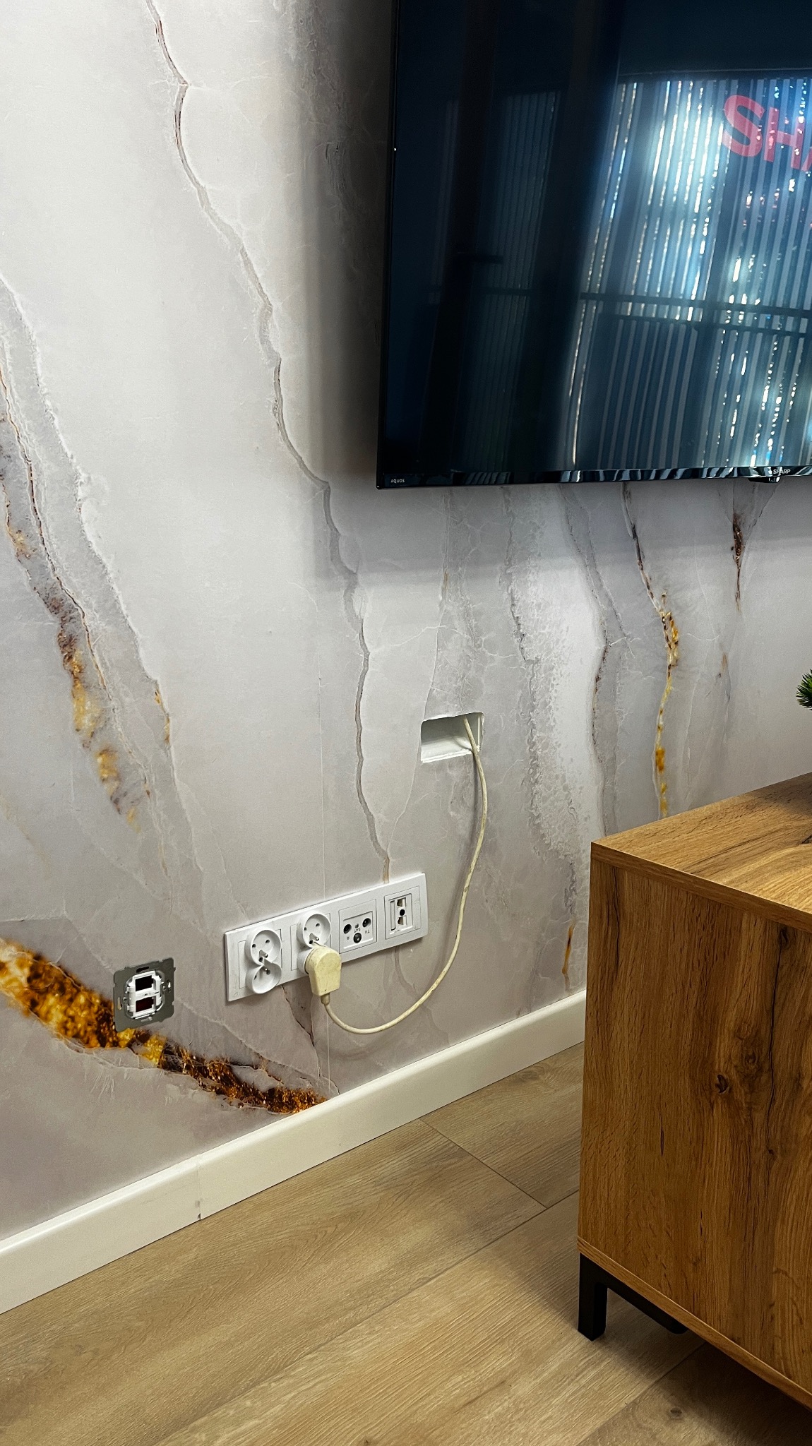 Świnoujście: Dekoracyjna ściana z efektem marmuru, z zamontowanym telewizorem i gniazdkami elektrycznymi, jedno z nich z wtyczką, oraz otwór na kable. Widoczny fragment drewnianej komody.