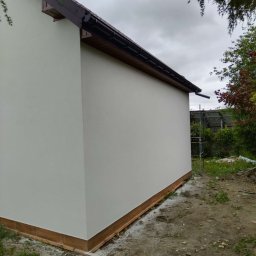 SOLID Adam Adamus - Świeżo otynkowana, biała ściana budynku z brązowym dachem i rynną. Widoczne rusztowanie z tyłu, zieleń i pochmurne niebo. Ukończony etap prac elewacyjnych.