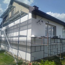 SOLID Adam Adamus - Dom w trakcie ocieplania, widoczne rusztowania i biały styropian na elewacji. Częściowo ocieplona ściana, z zamontowaną anteną. Prace w toku, słoneczny dzień.