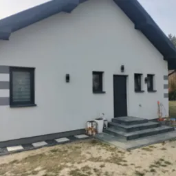 Elewacja domu jednorodzinnego w odcieniach szarości z nowoczesnym oświetleniem zewnętrznym. Widoczne schody wejściowe i fragment ogrodu.