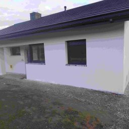 SOLID Adam Adamus - Otynkowany dom jednorodzinny z ciemnym dachem i oknami. Widoczny fragment elewacji bocznej i otoczenie domu. Na ziemi stoi wiadro z farbą.