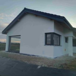 Nowoczesny dom jednorodzinny z białymi ścianami i ciemną dachówką, widoczny z zewnątrz, z dużym oknem i elementami wykończeniowymi w kolorze antracytowym.