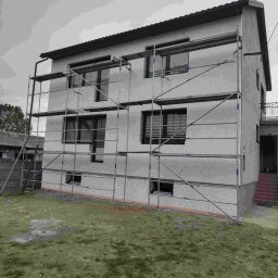 Dom jednorodzinny w trakcie ocieplania, z widocznymi rusztowaniami i szarym tynkiem. Okna z roletami. Widok z poziomu gruntu na elewację.